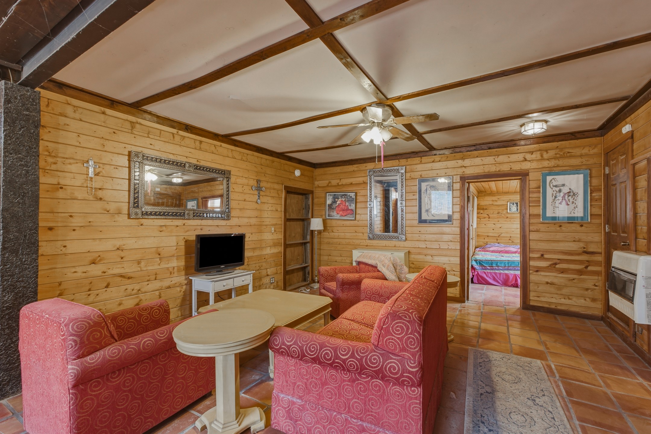 Wood-paneled living room with red patterned sofas, terracotta tile, and an open doorway to a bedroom.