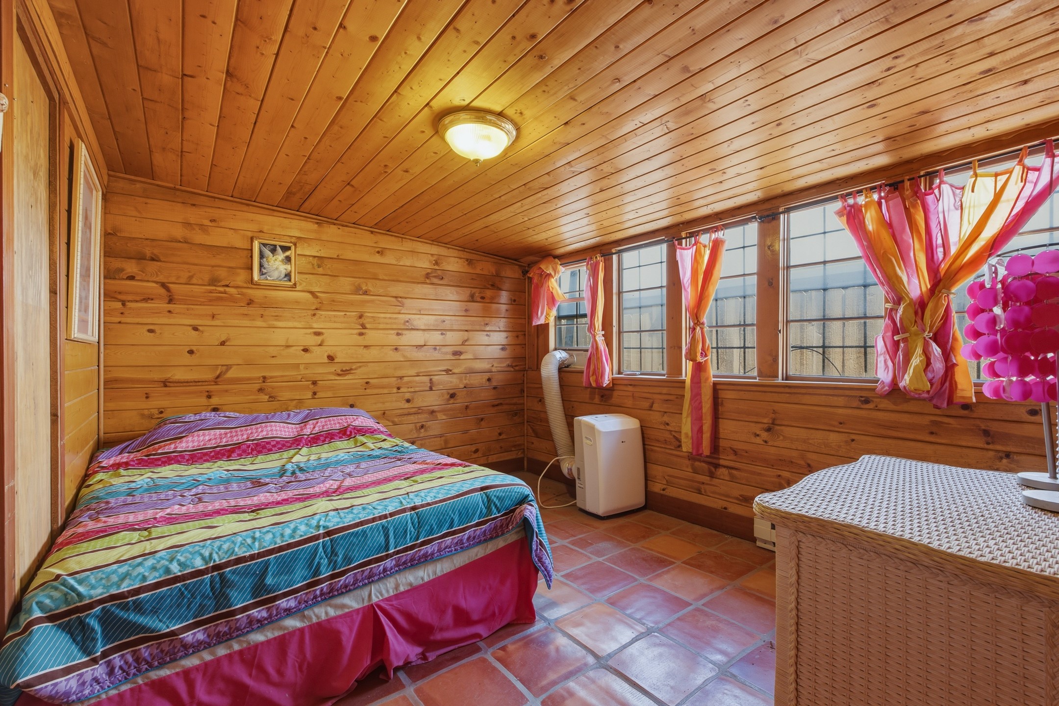 Bedroom with wood-paneled walls, colorful striped bedding, and bright curtains over a row of windows.