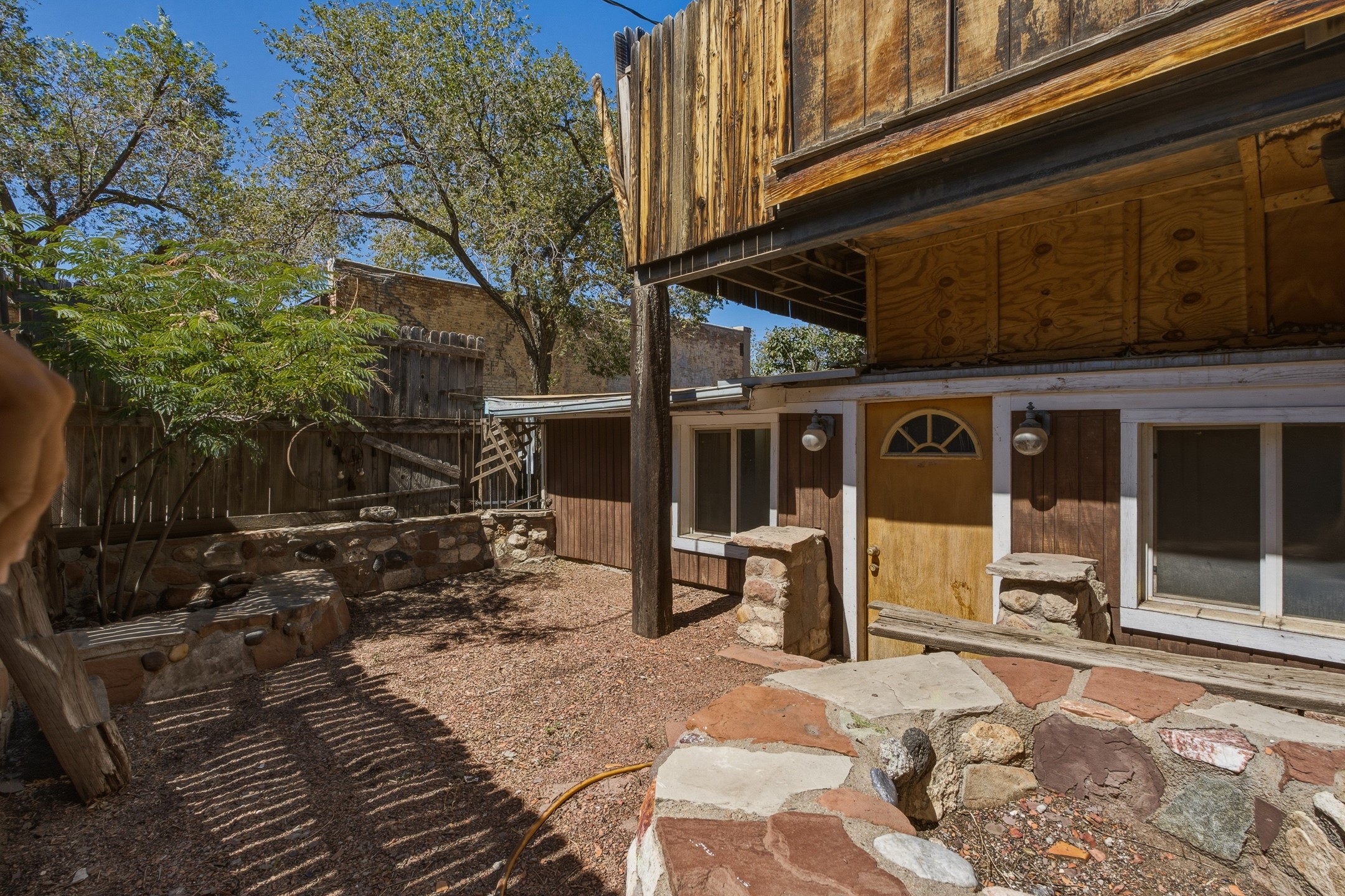 Home courtyard with a wooden upper deck, yellow entry door, stone seating walls, and mature trees.