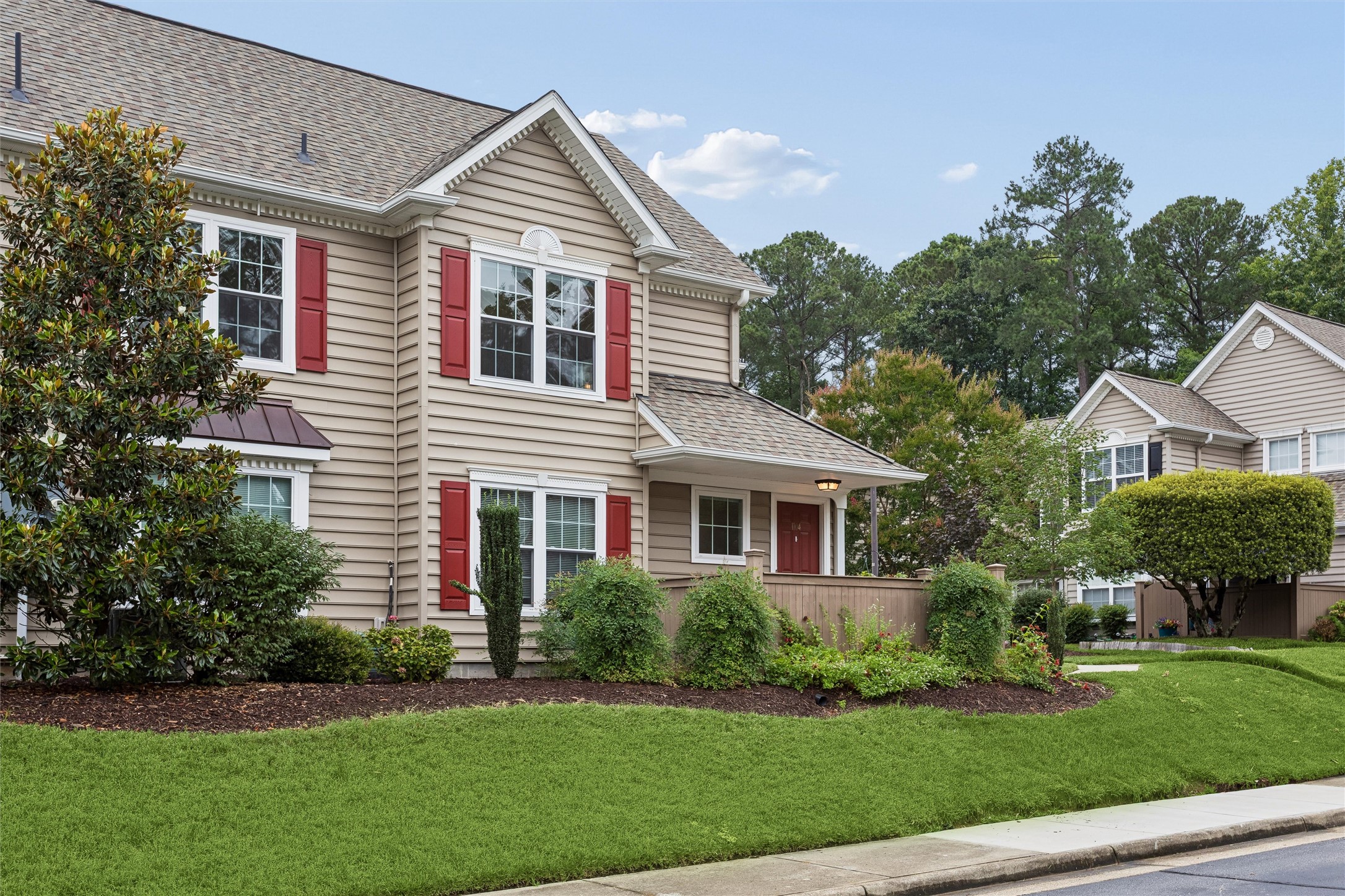 Two-story beige house with red shutters, a covered porch, and landscaped front lawn.