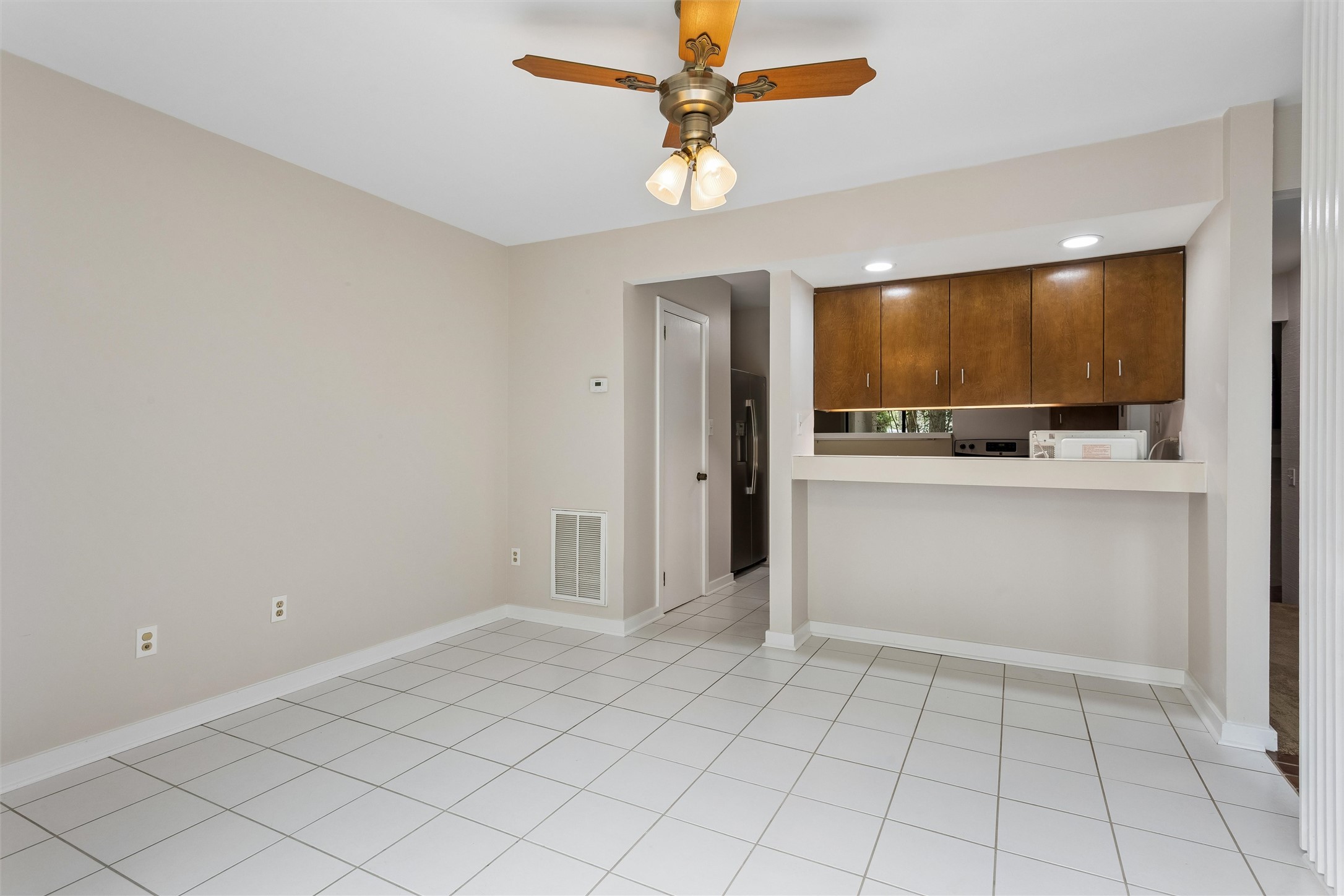 Empty tiled dining area beside a kitchen with dark wood cabinets and a ceiling fan.