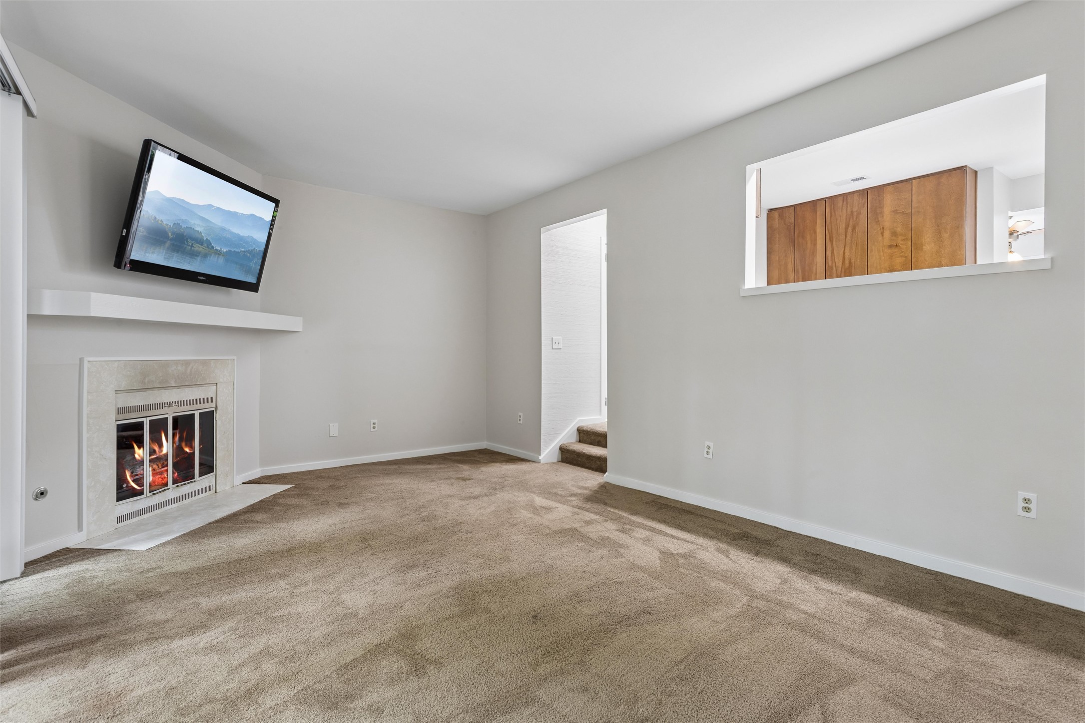 Unfurnished living room with a gas fireplace, wall-mounted television, beige carpet, and pass-through to wooden cabinets.