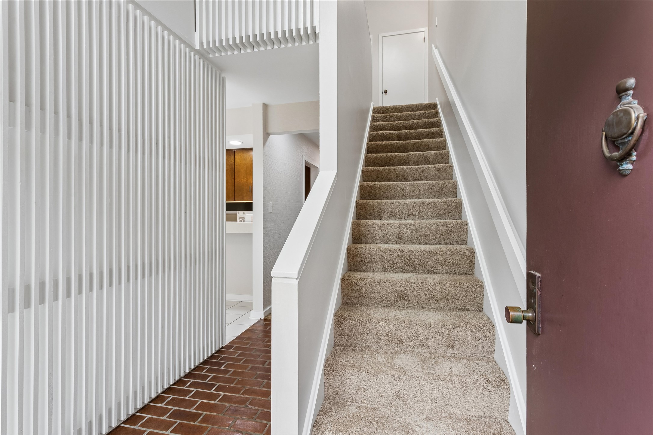 Entryway staircase with beige carpeting, a tall white slatted divider, and brick tile flooring.