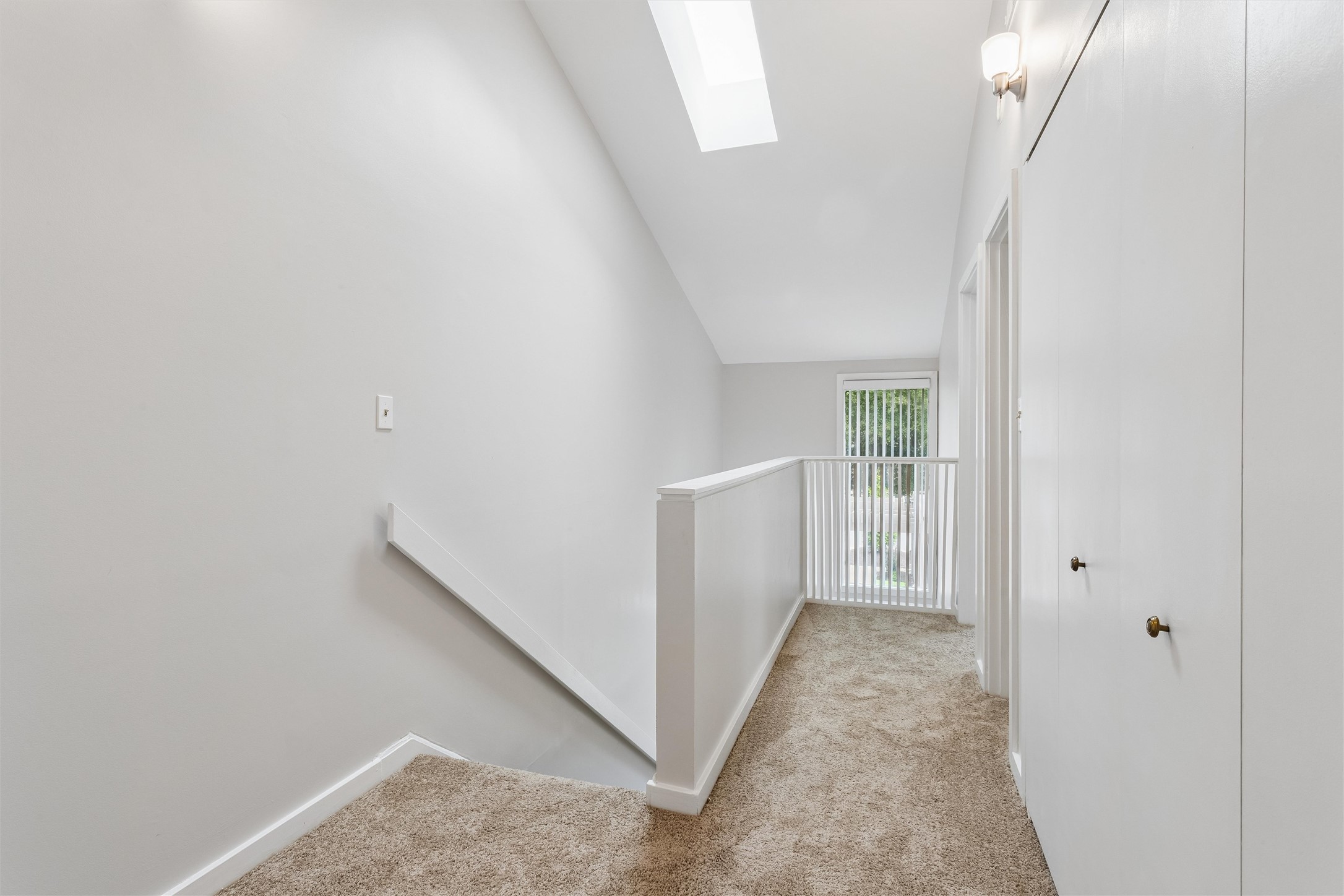 Carpeted upstairs landing with white railing, skylight, and window with vertical blinds.