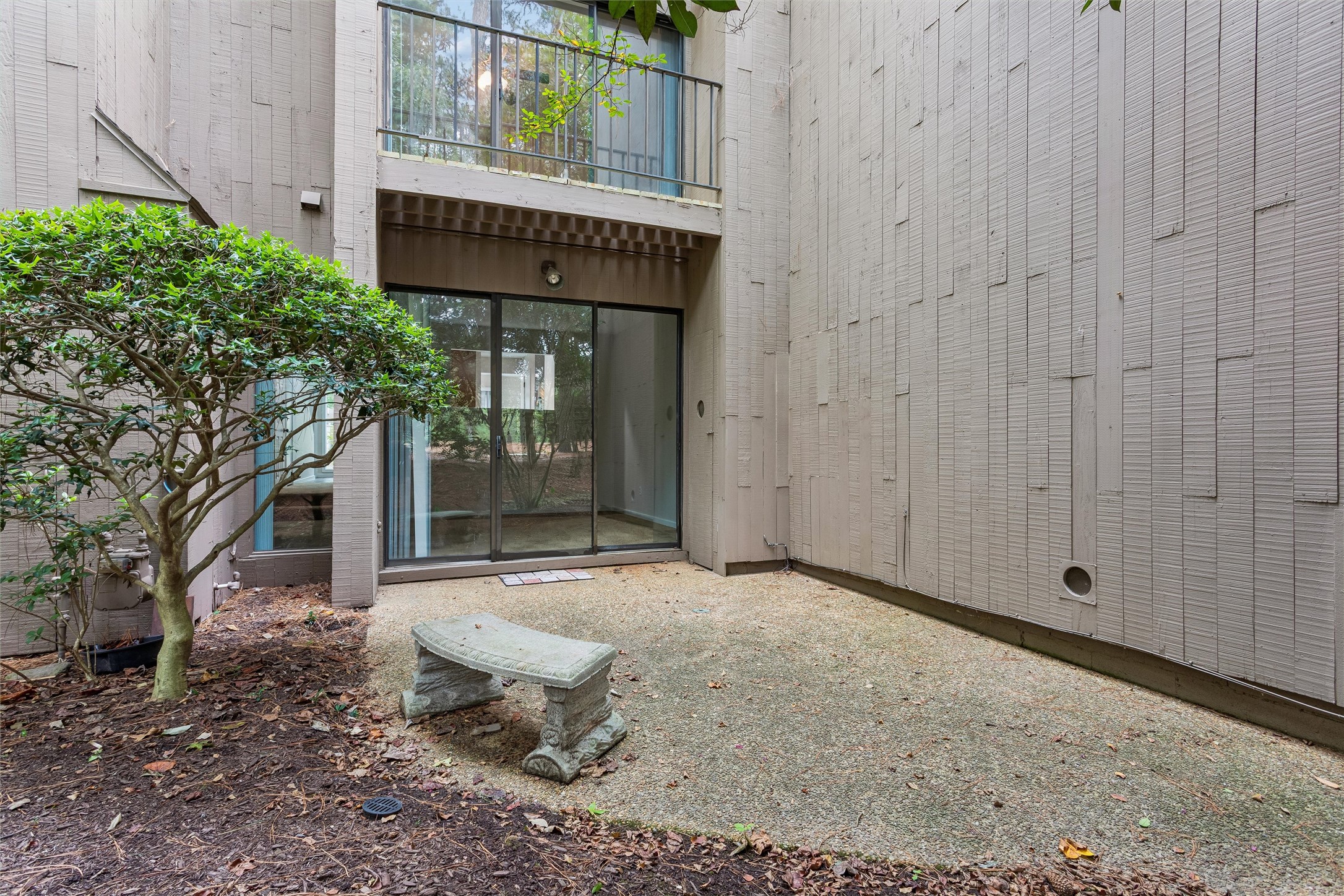 Courtyard patio with a stone bench, leafy tree, sliding glass doors, and upper balcony.