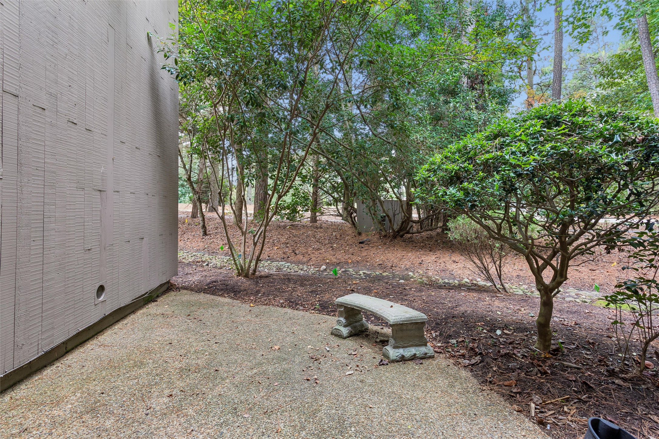 Gravel patio beside a house, with a stone bench among leafy trees and shrubs.