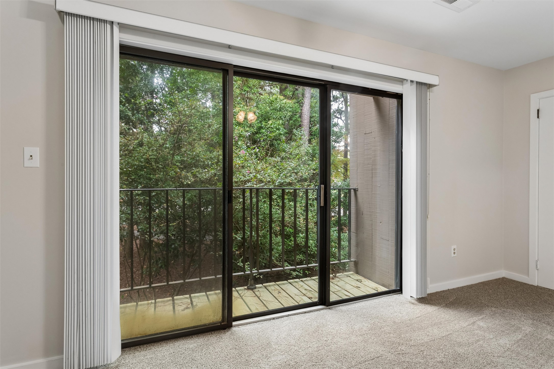 Empty carpeted room with sliding glass doors overlooking a wooded balcony.