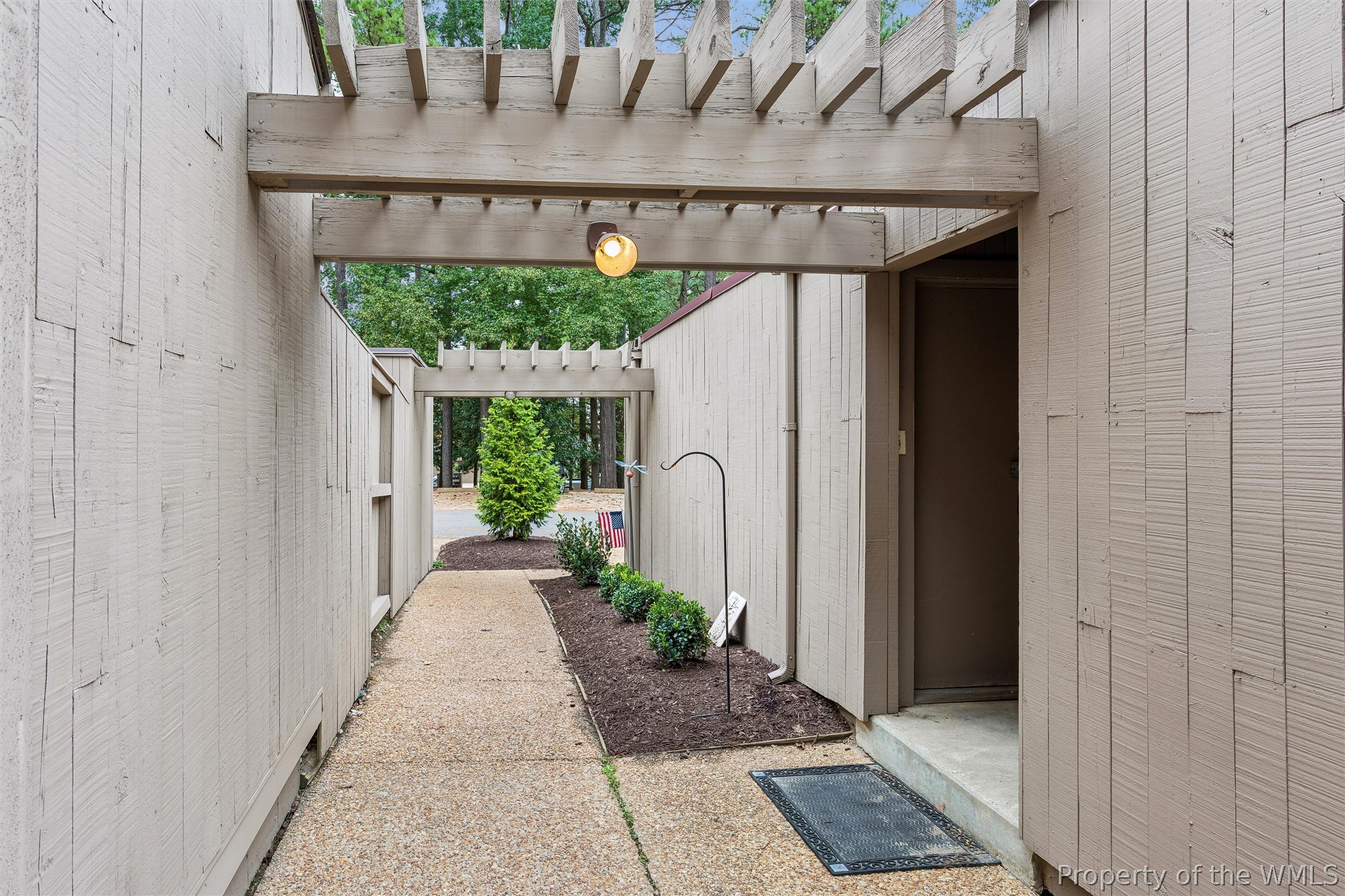 Home entrance beneath a wooden pergola, with a pebble-finish walkway and landscaped planting beds.