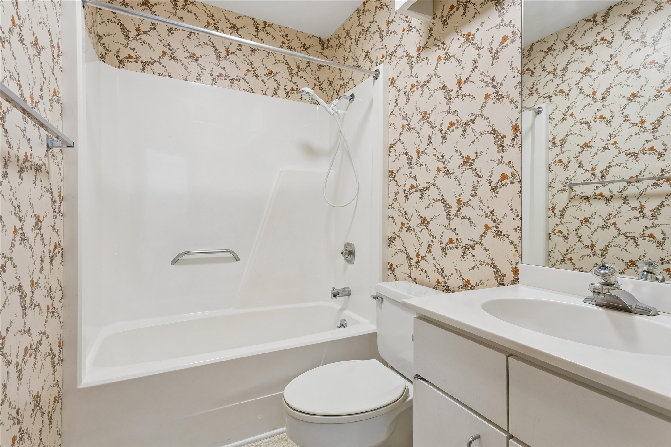 Bathroom with a white tub-shower, floral wallpaper, toilet, and sink vanity.
