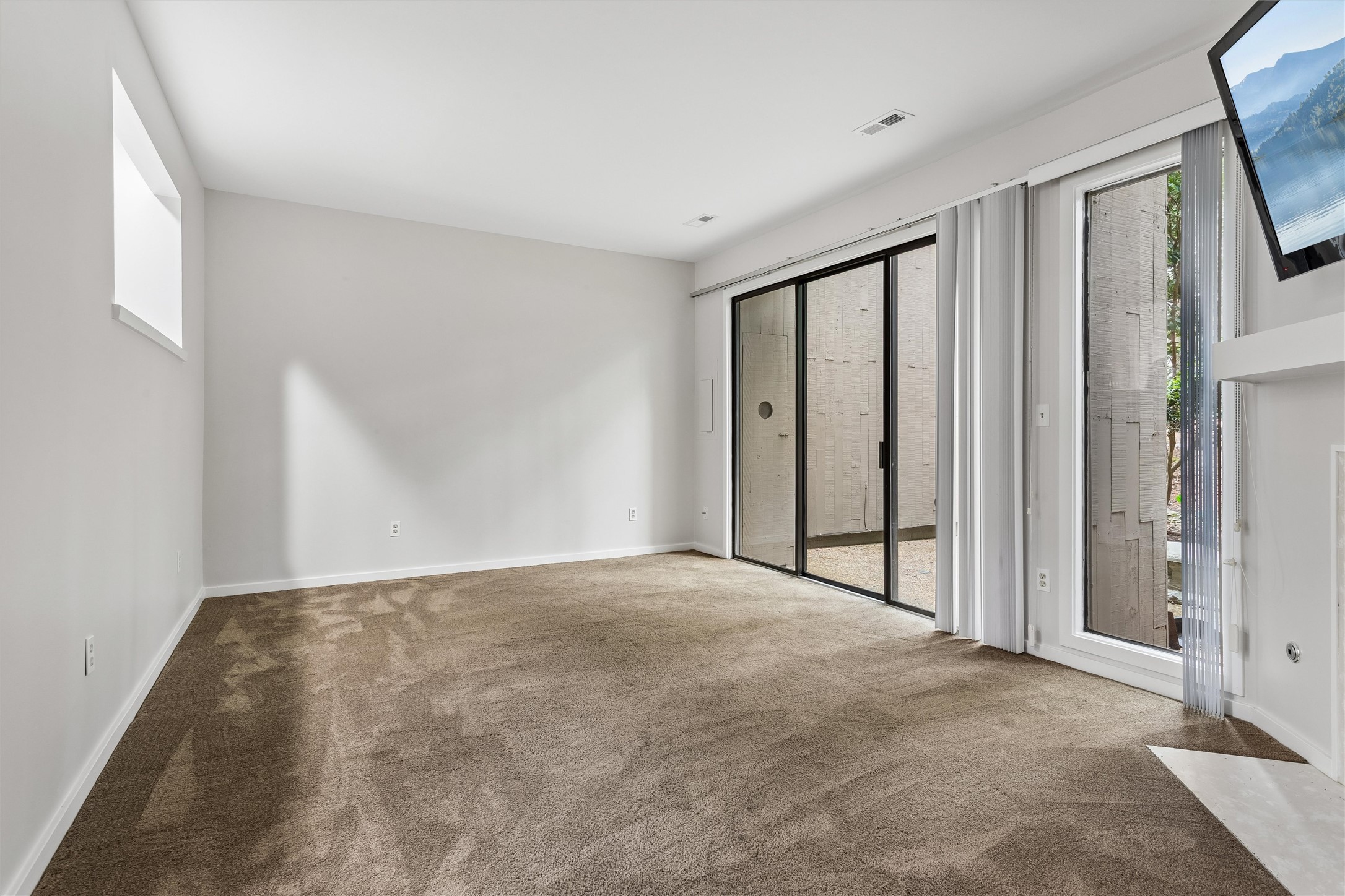 Vacant living room with beige carpet and sliding glass doors with vertical blinds.