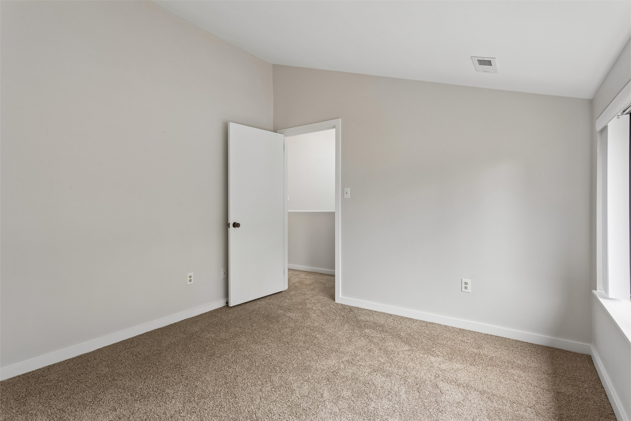 Empty carpeted bedroom with pale walls, an open door, and a window.