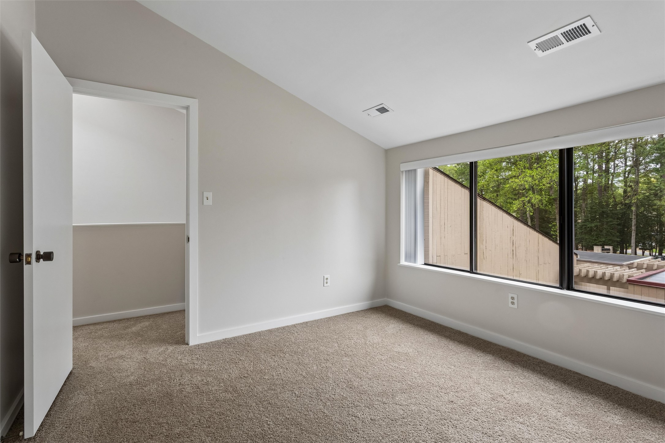 Empty carpeted room with a wide window overlooking trees and nearby rooftops.