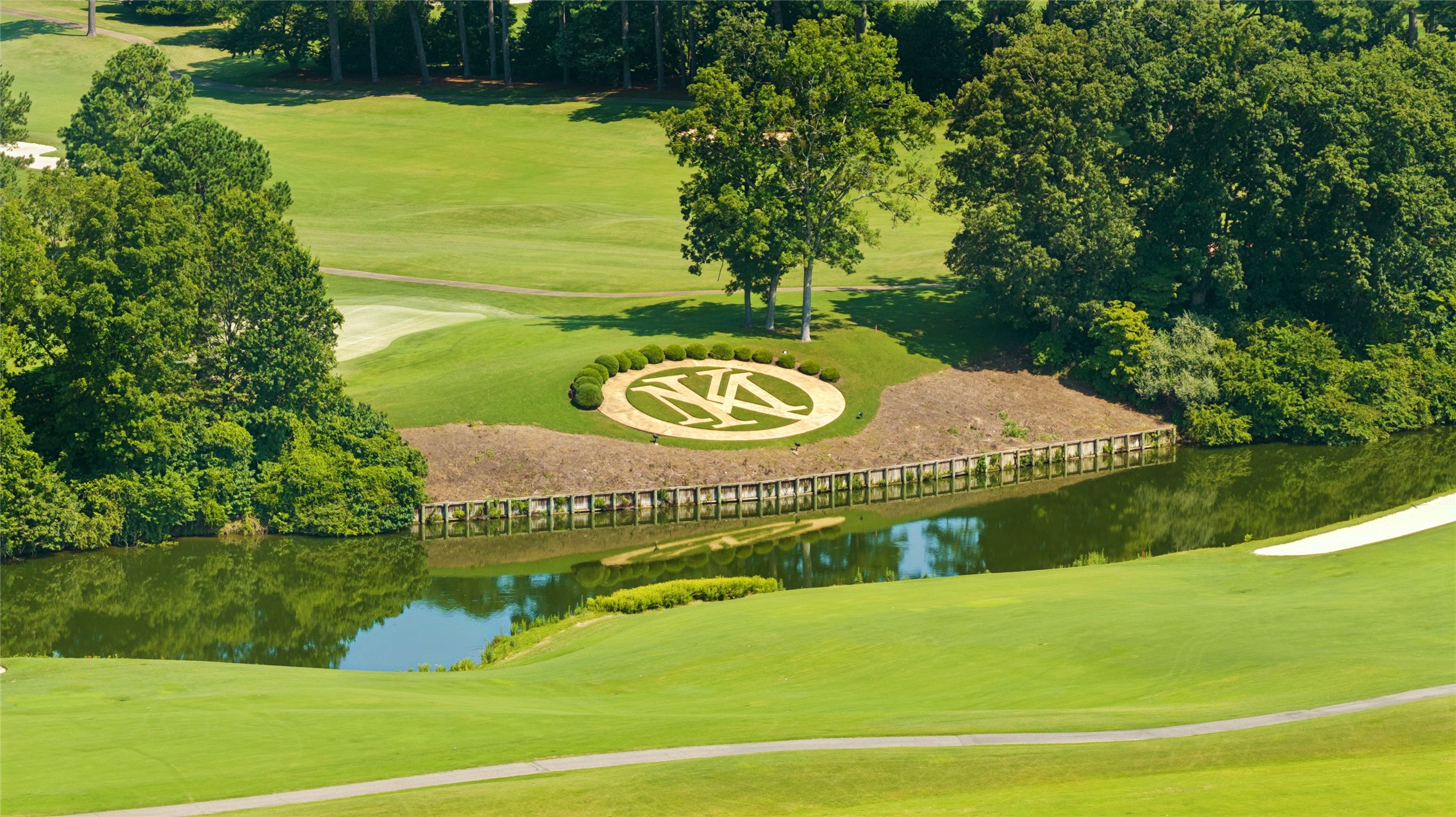 Aerial view of a golf course with a pond, tree-lined grounds, and a circular landscaped emblem.