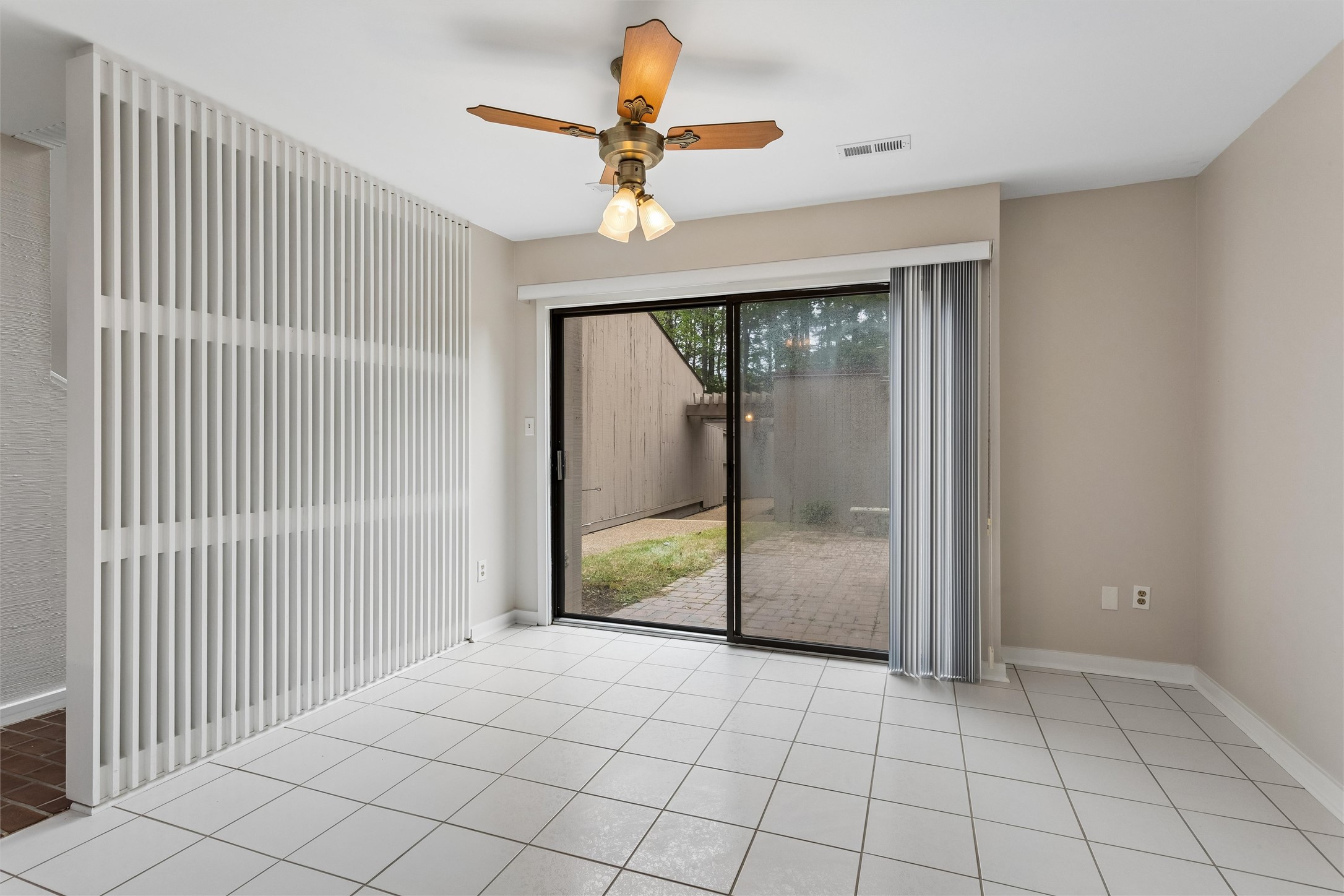 Empty tiled room with a white slat divider and sliding glass doors to a paved patio.