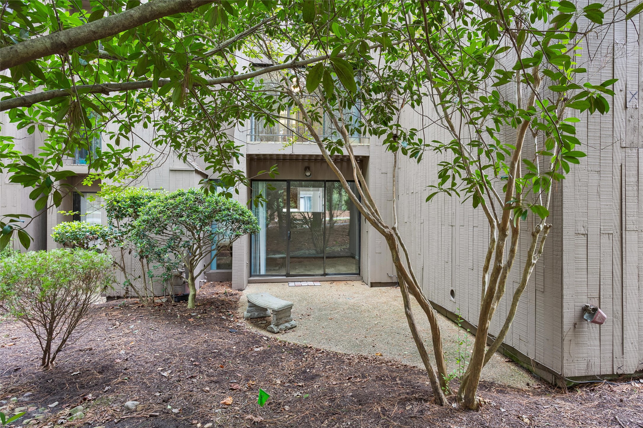 Residential courtyard with a gravel patio, stone bench, sliding glass doors, and mature trees.