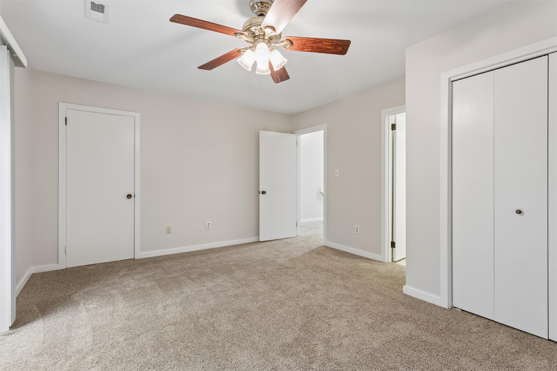 Empty bedroom with beige carpet, two closets, and a lit ceiling fan.