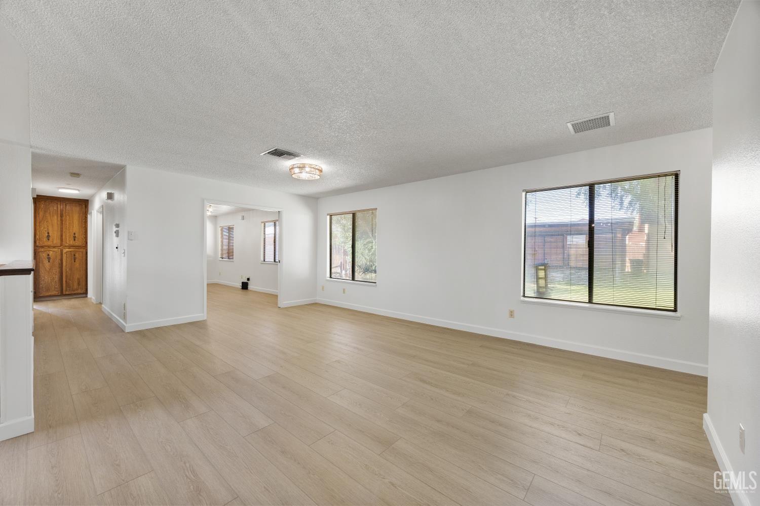Vacant living room with light plank flooring, white walls, and large windows with blinds.