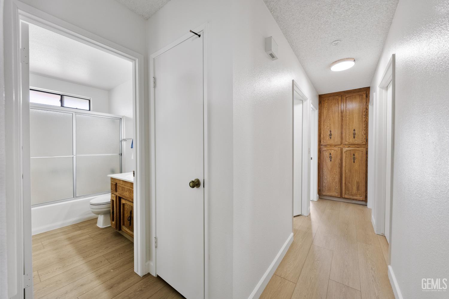 Hallway with light wood-look flooring, a bathroom doorway, and a tall wood cabinet at the far end.
