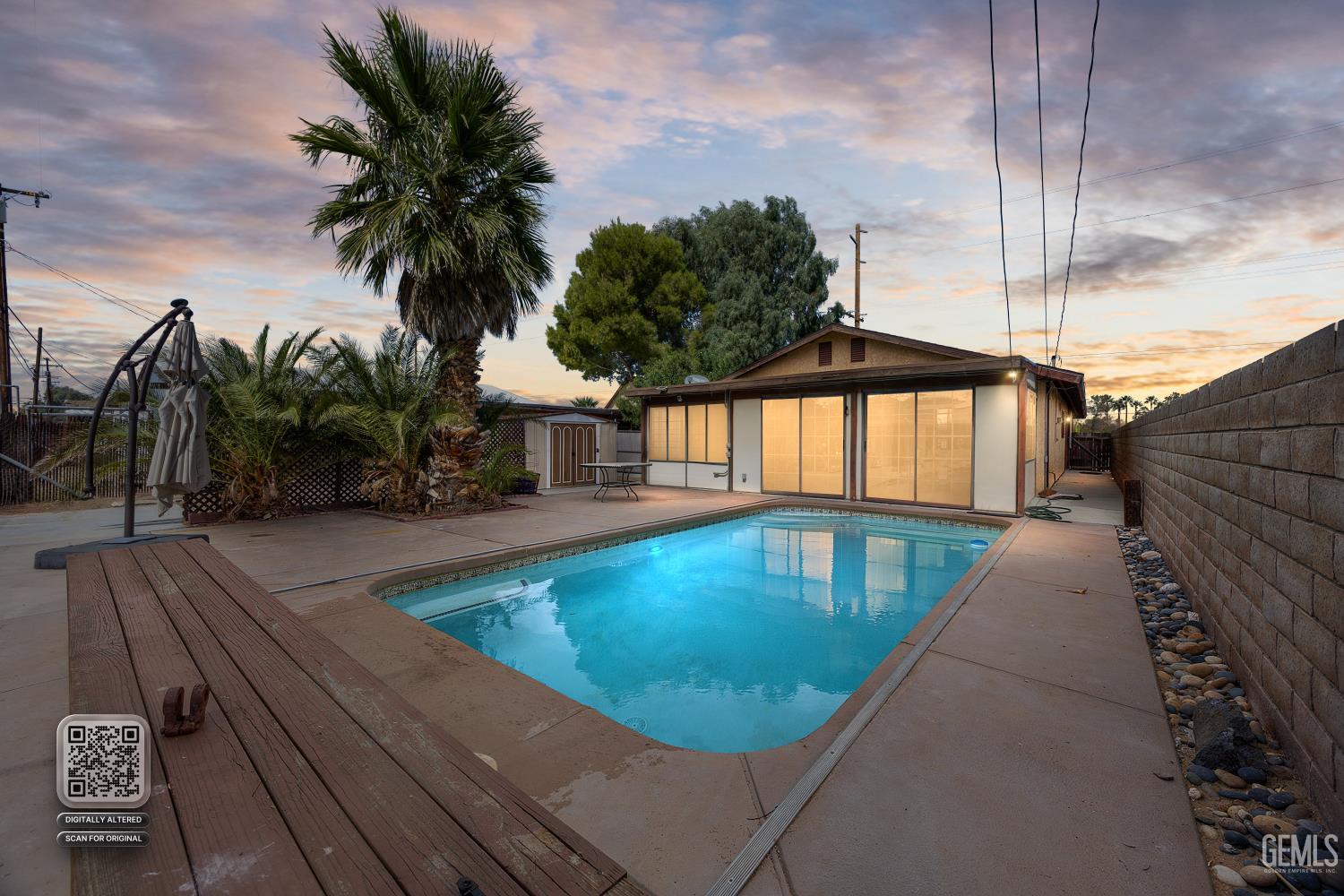 Backyard pool and patio beside a single-story house at dusk, with palm trees and illuminated windows.