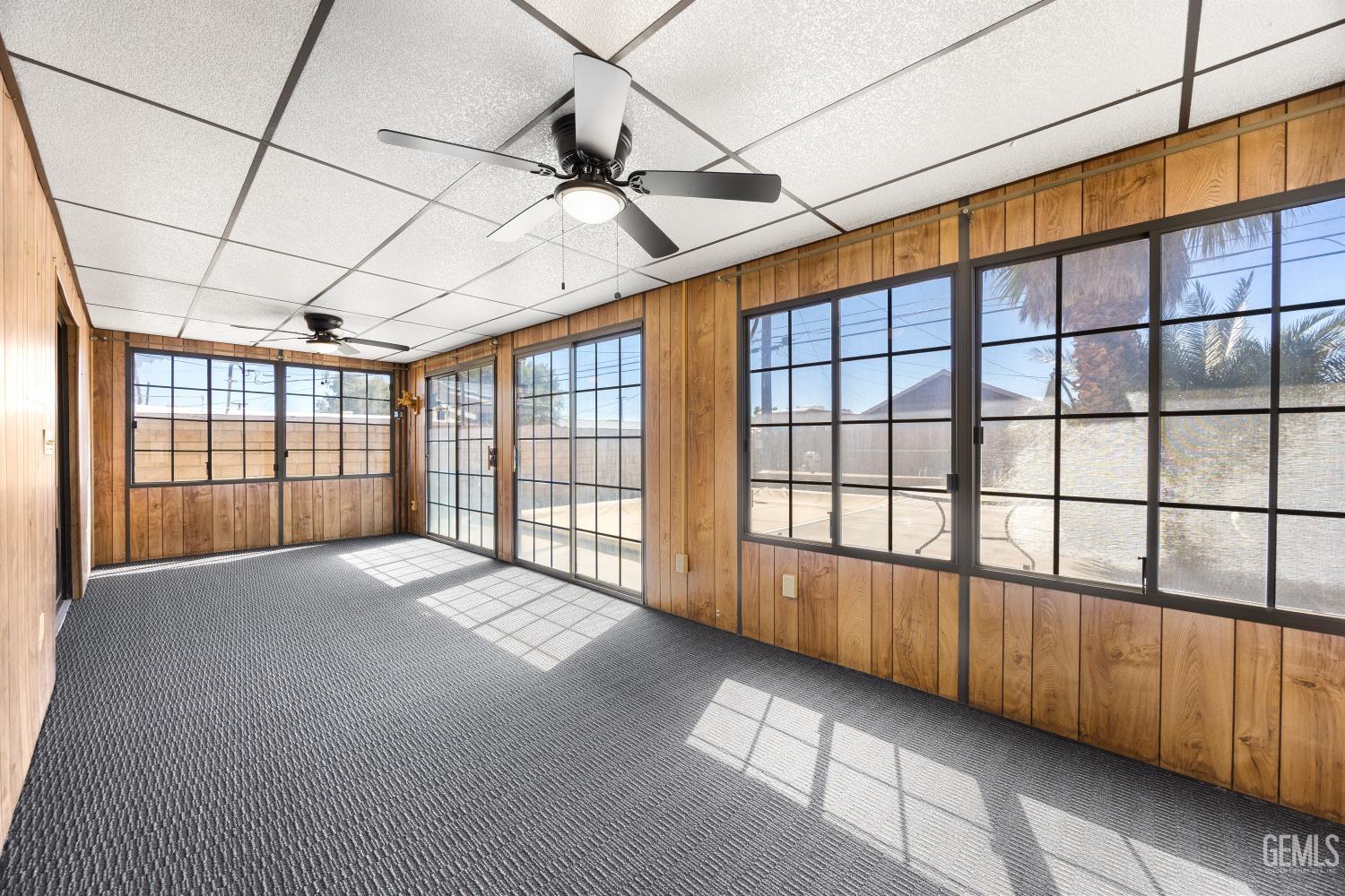 Enclosed sunroom with wood-paneled walls, carpet, ceiling fans, and broad windows overlooking a patio.