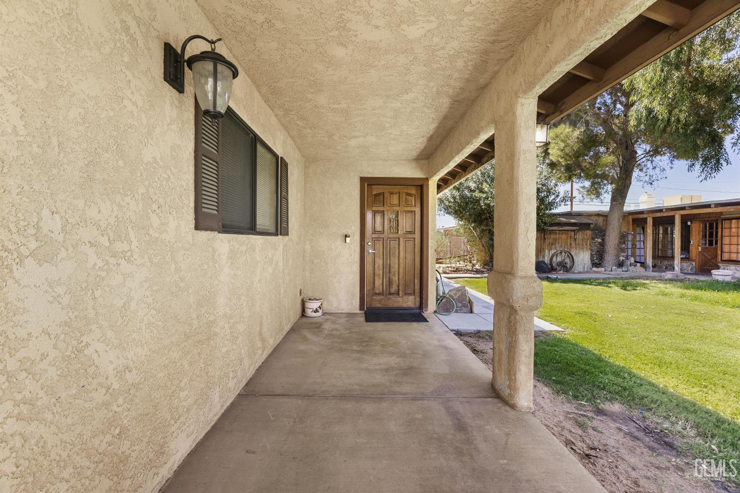 Covered stucco porch with a wooden front door and a view of the lawn and backyard outbuildings.