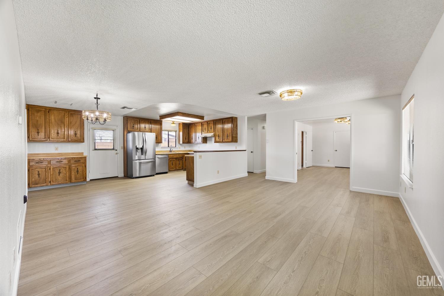 Open living area and kitchen with wood cabinetry, stainless steel refrigerator, and light wood-look flooring.