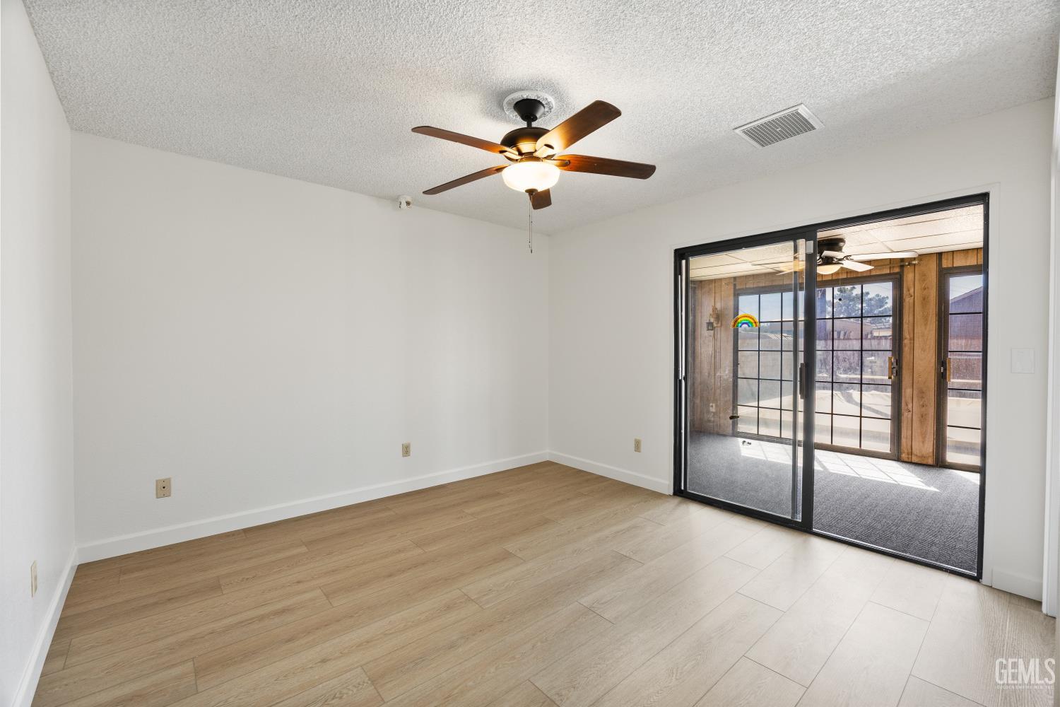 Empty room with wood-look tile floors, ceiling fan, and sliding doors to a sunlit enclosed patio.