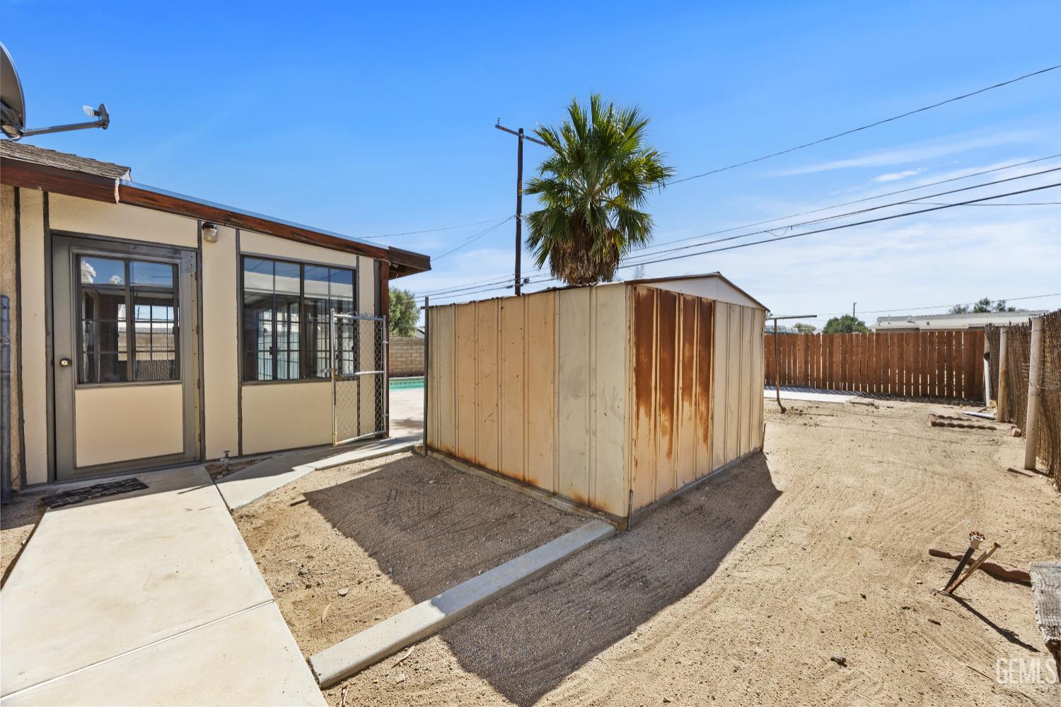 Sandy backyard with a weathered metal shed, concrete walkway, wooden fence, and a glimpse of the pool.