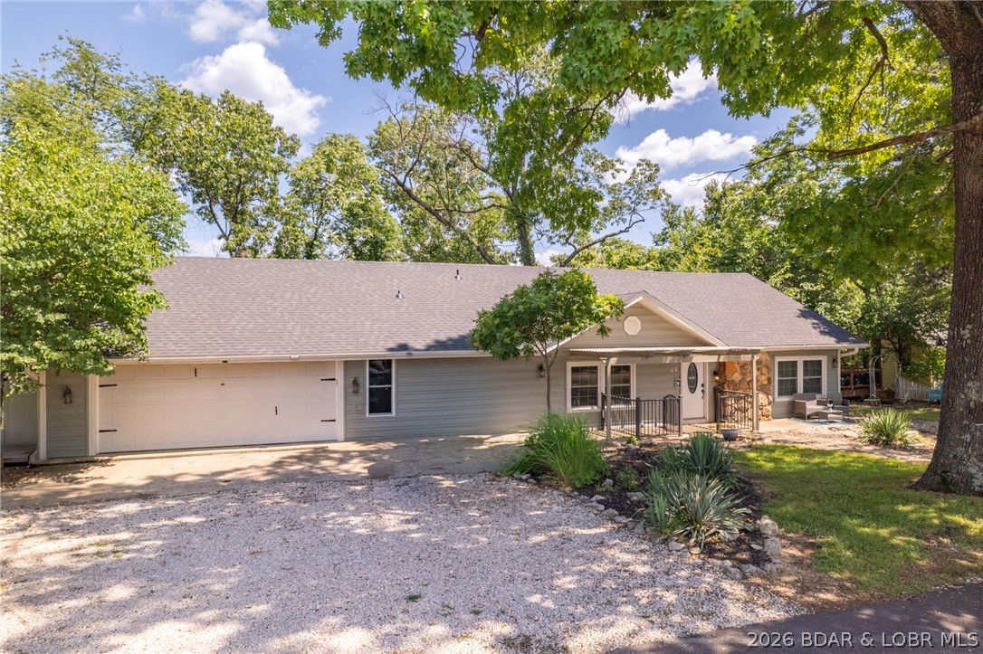 Single-story gray home with a two-car garage, stone-accented entry, and gravel driveway beneath mature trees.
