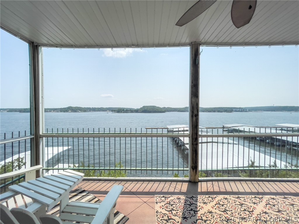 Covered balcony overlooking a lake and boat docks, with white lounge chairs and a ceiling fan.