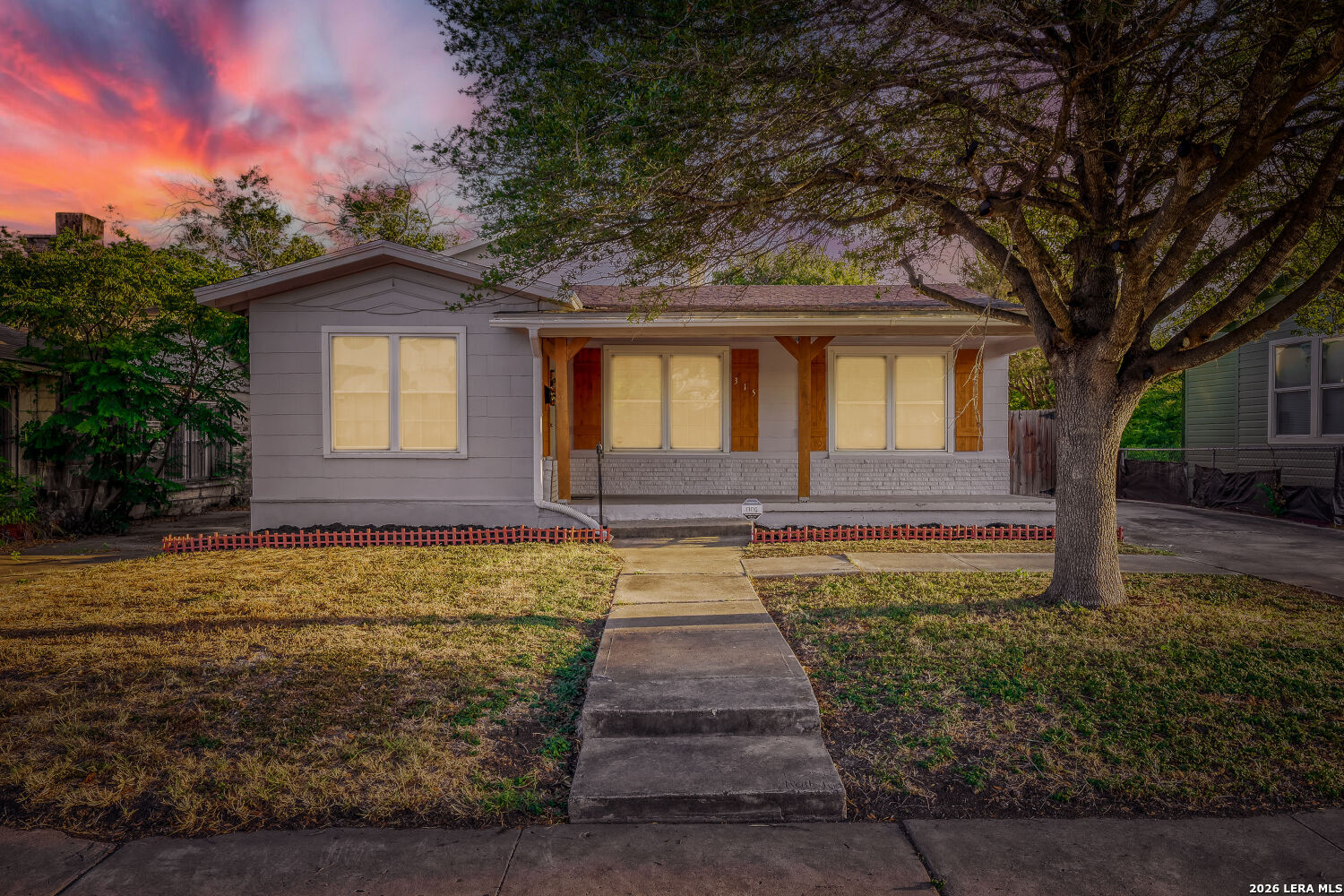 Single-story gray house with a covered porch and large front-yard tree at dusk.