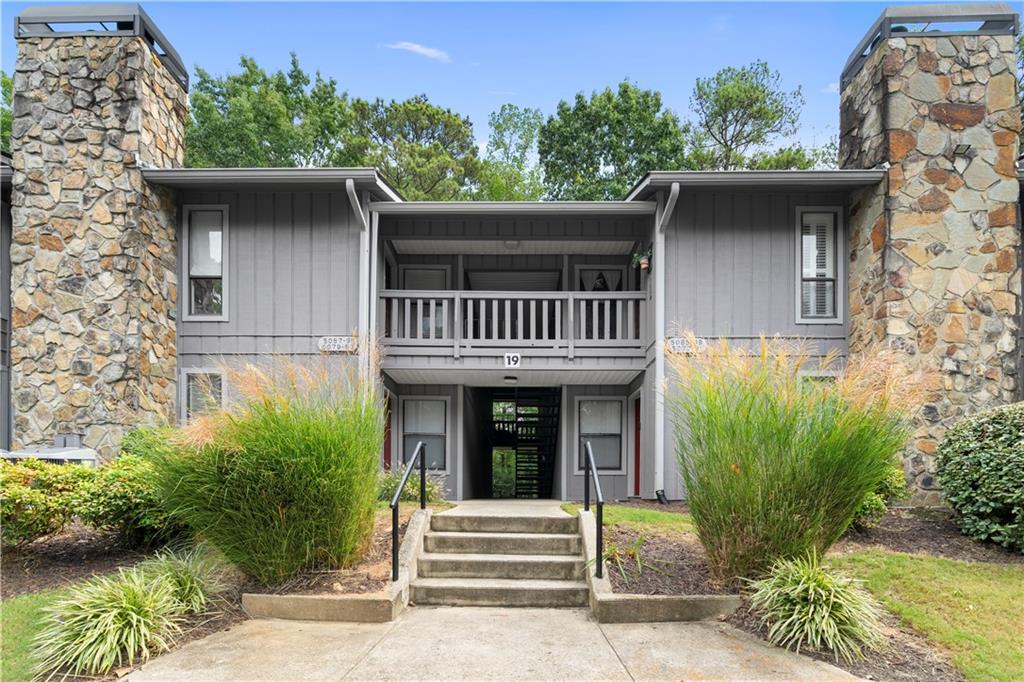 Gray residential building entrance with a covered balcony, stone columns, and landscaped steps.