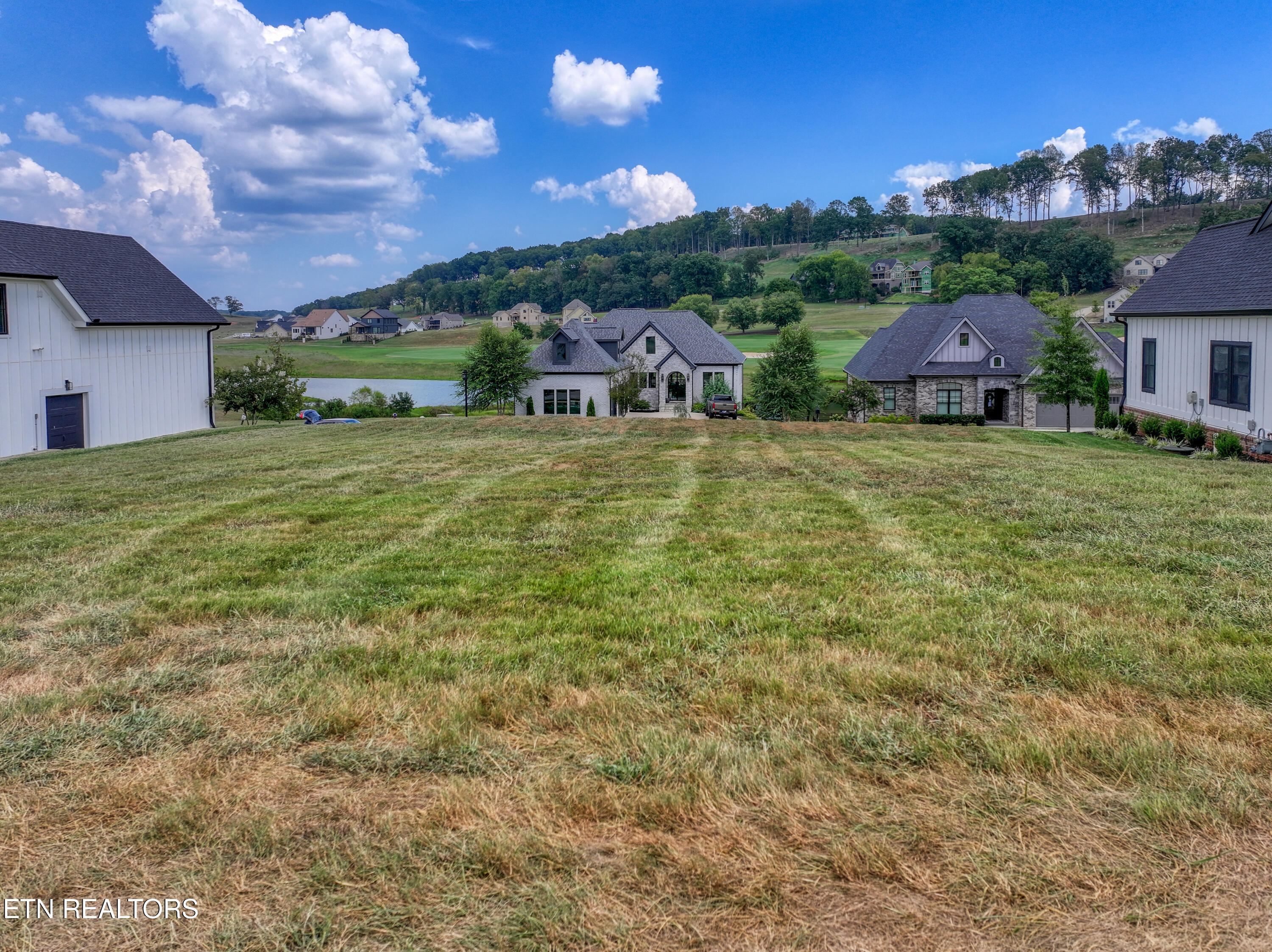 Grassy lot sloping toward a lake, with neighboring homes and wooded hills beyond.