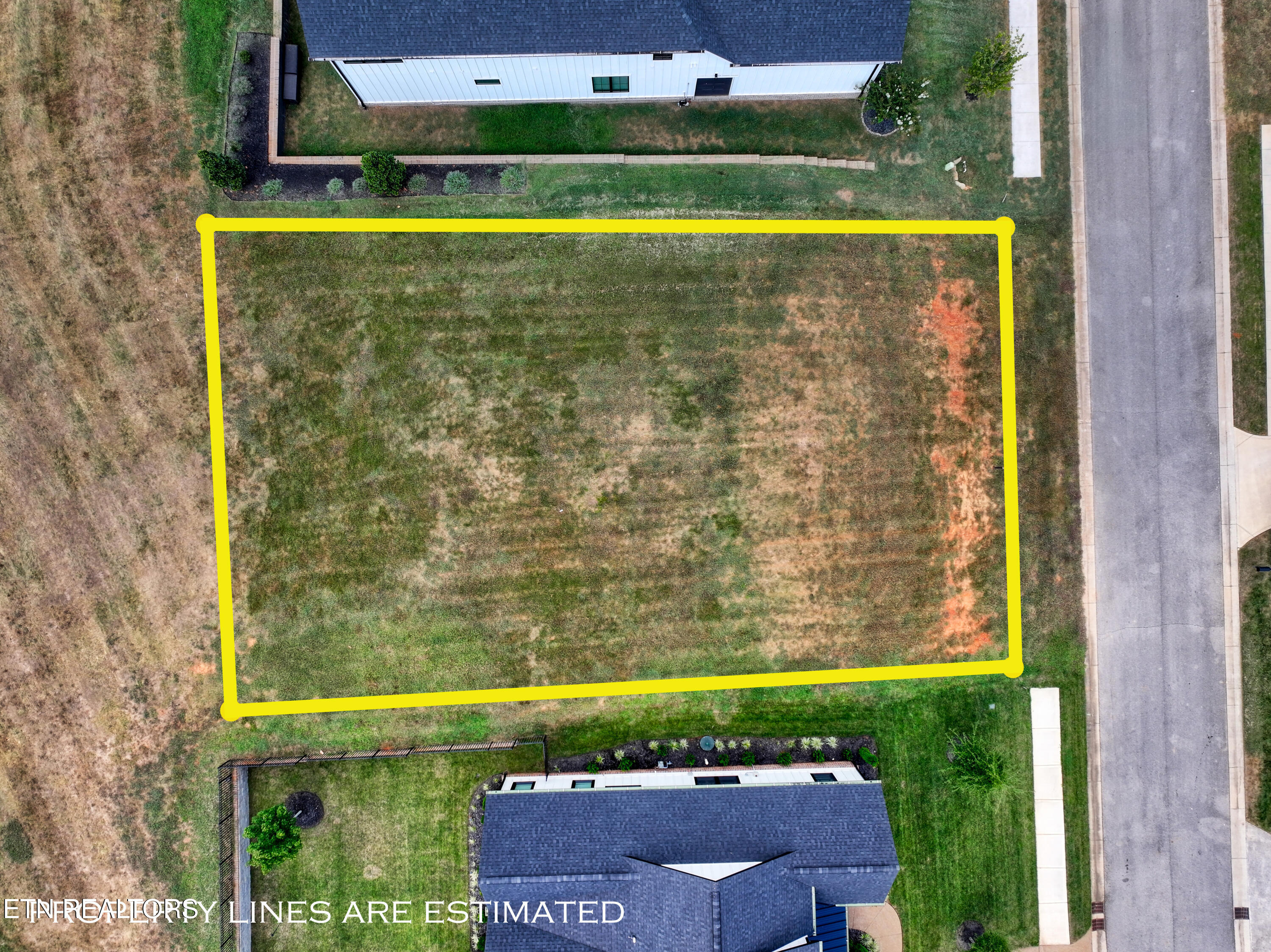 Aerial view of a grassy lot outlined in yellow between two homes, beside a road.
