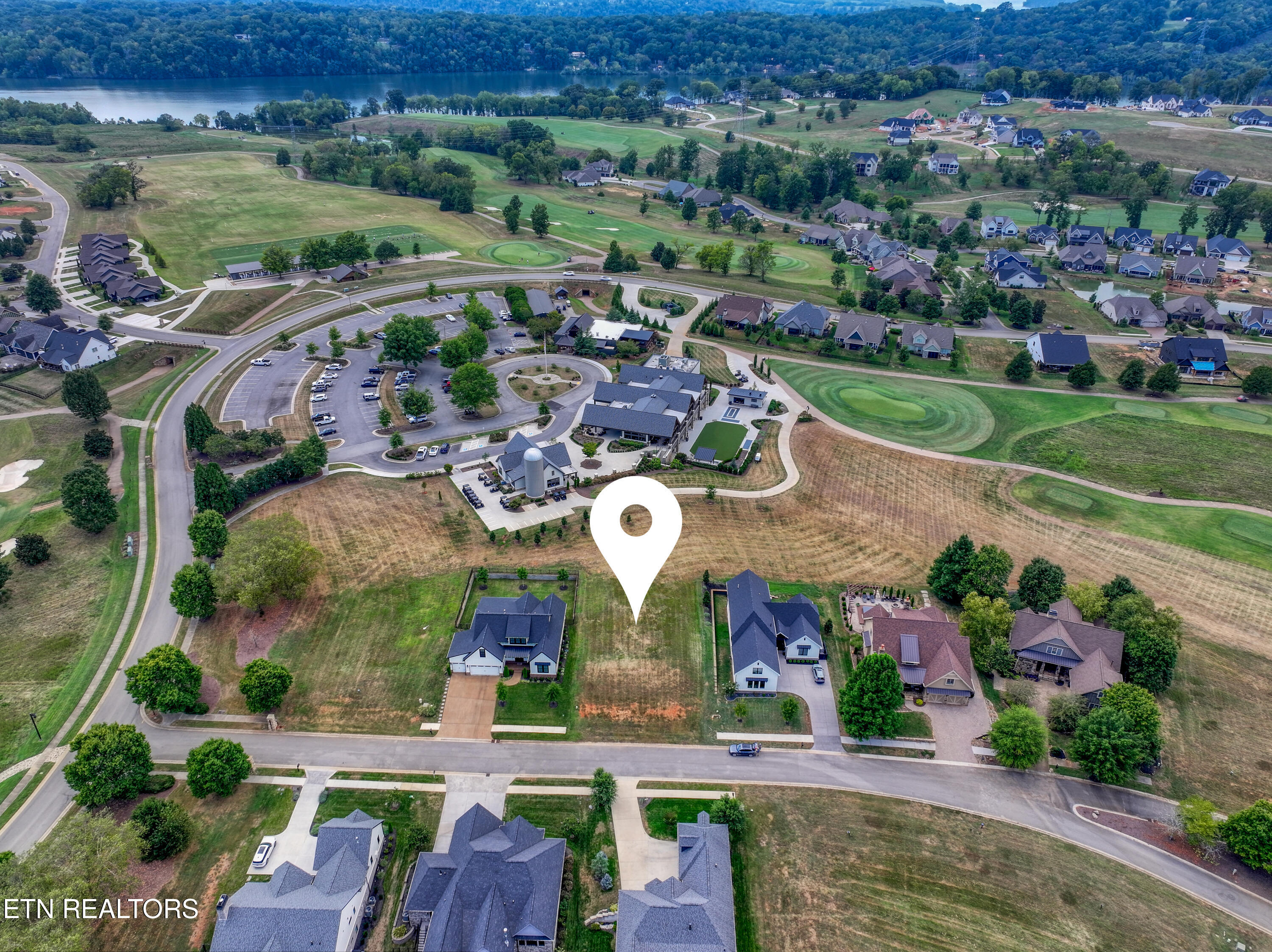 Aerial neighborhood view with the property marked by a pin, landscaped greens, and a lake in the distance.