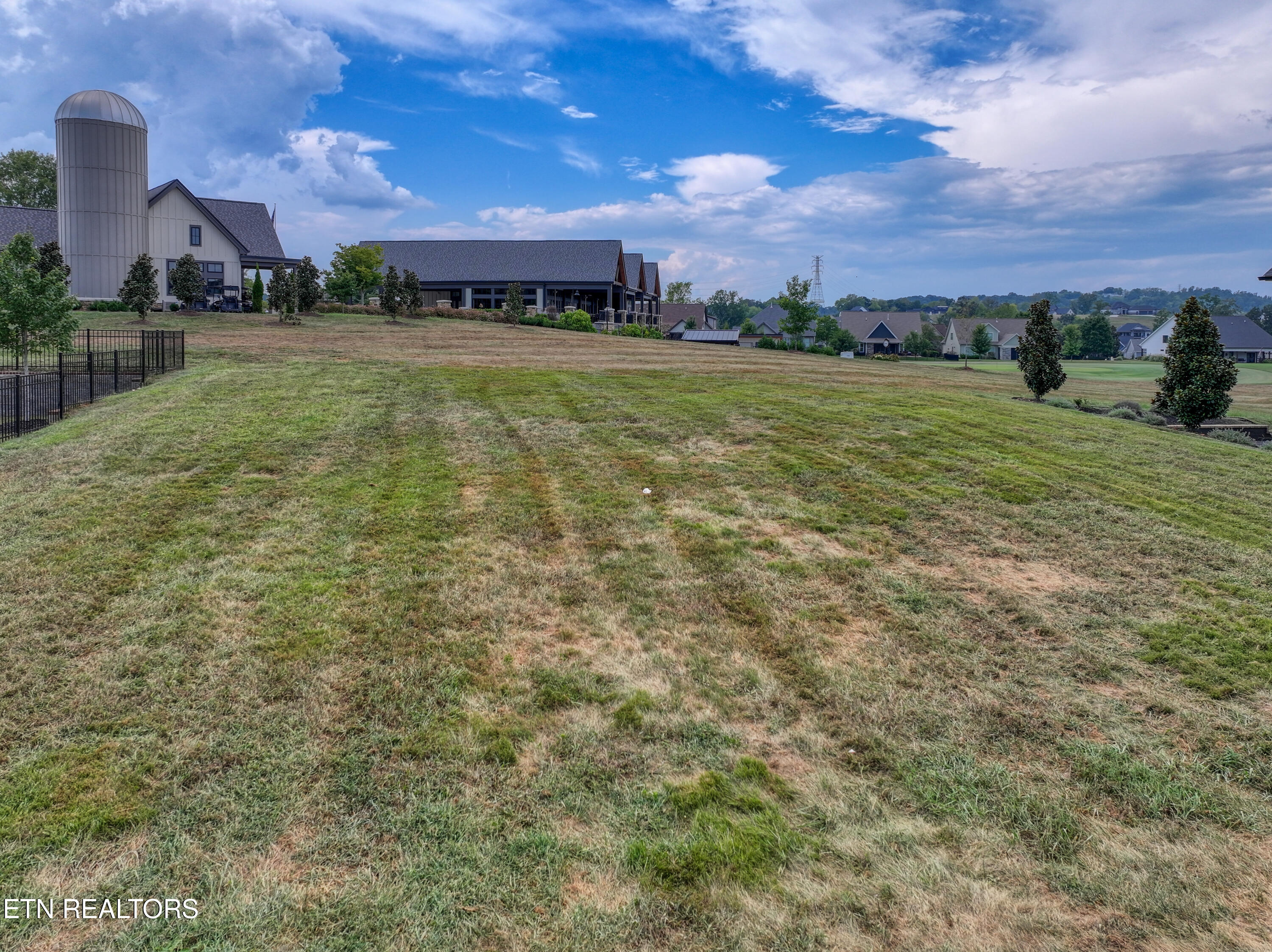 Sloping grassy lot with a barn-style building, tall silo, and neighboring homes in the distance.