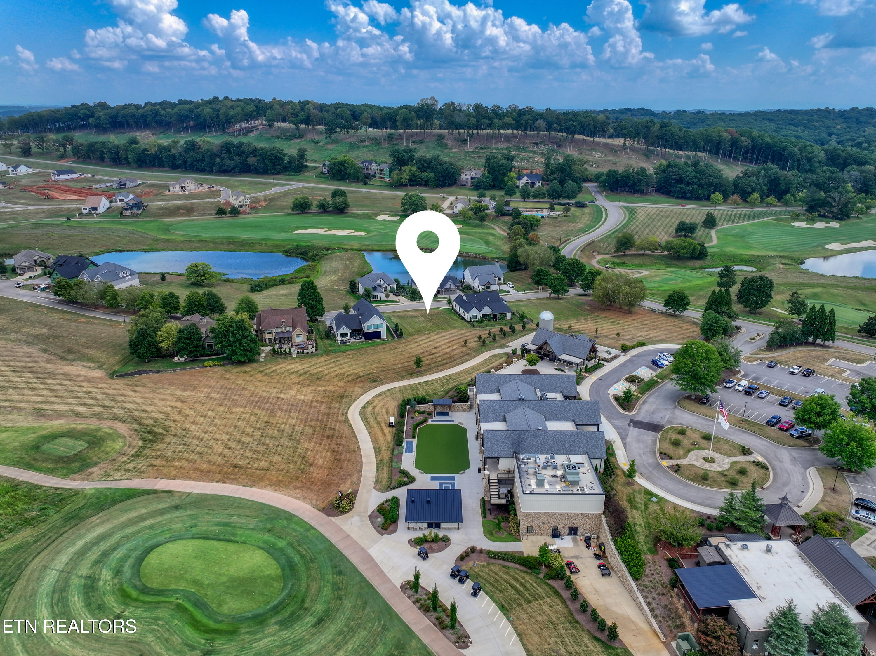 Aerial view of a home marked beside a golf course and pond, with wooded hills beyond.