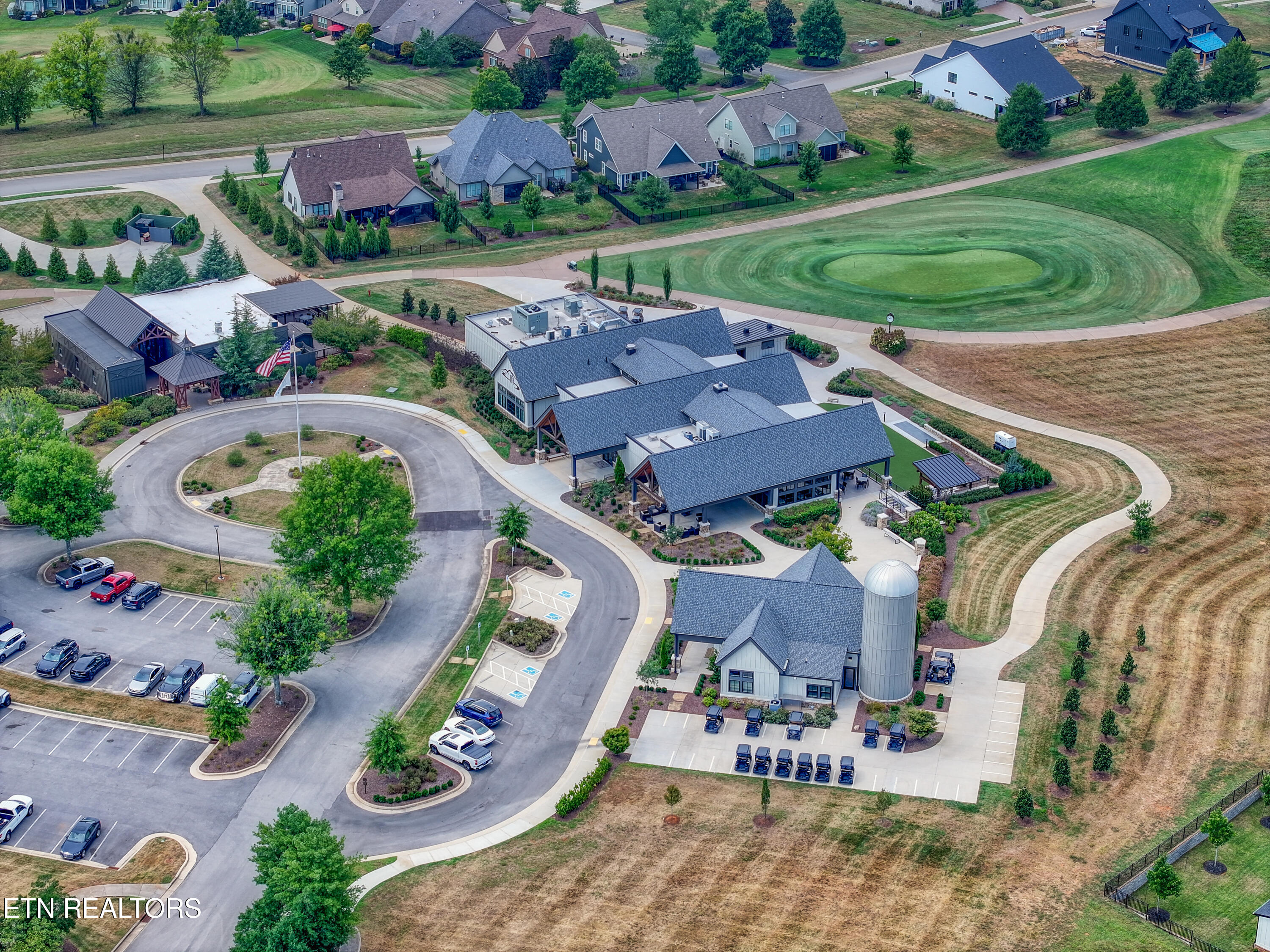 Aerial view of a neighborhood complex with blue-gray-roofed buildings, parking, nearby homes, and an oval lawn.