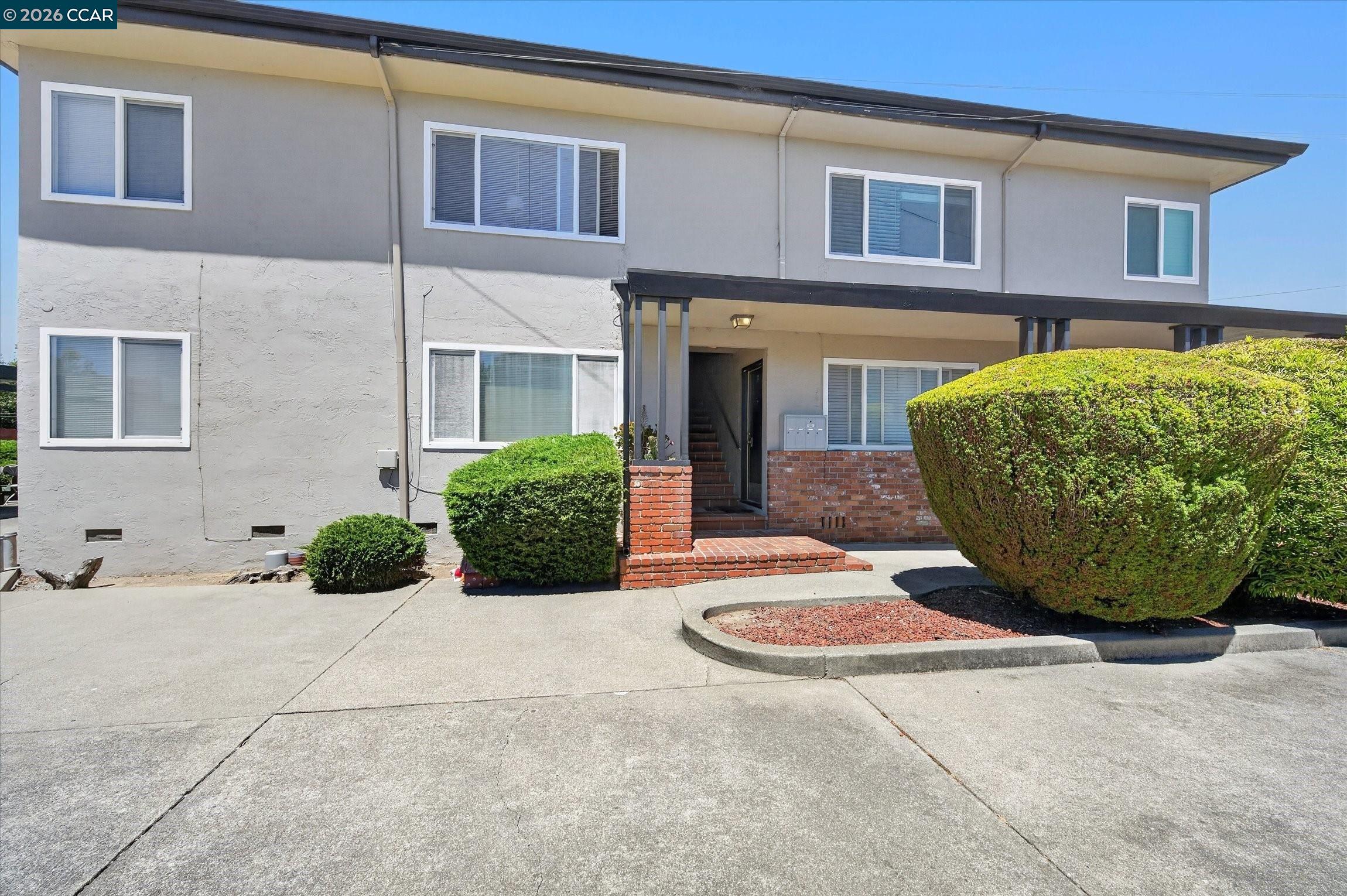 Two-story gray residential building with a covered brick entrance and large trimmed shrubs.