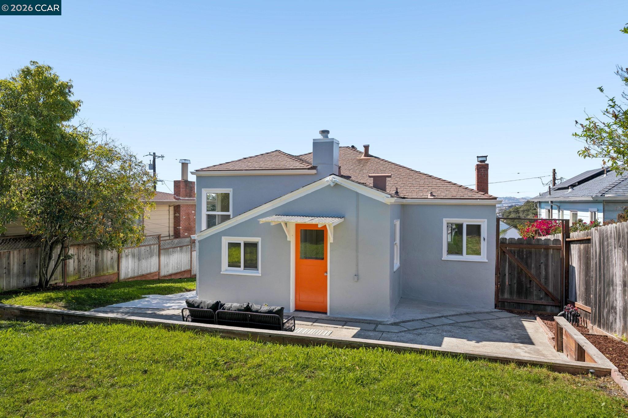 Rear of a gray house with a bright orange door, patio seating, and a fenced lawn.
