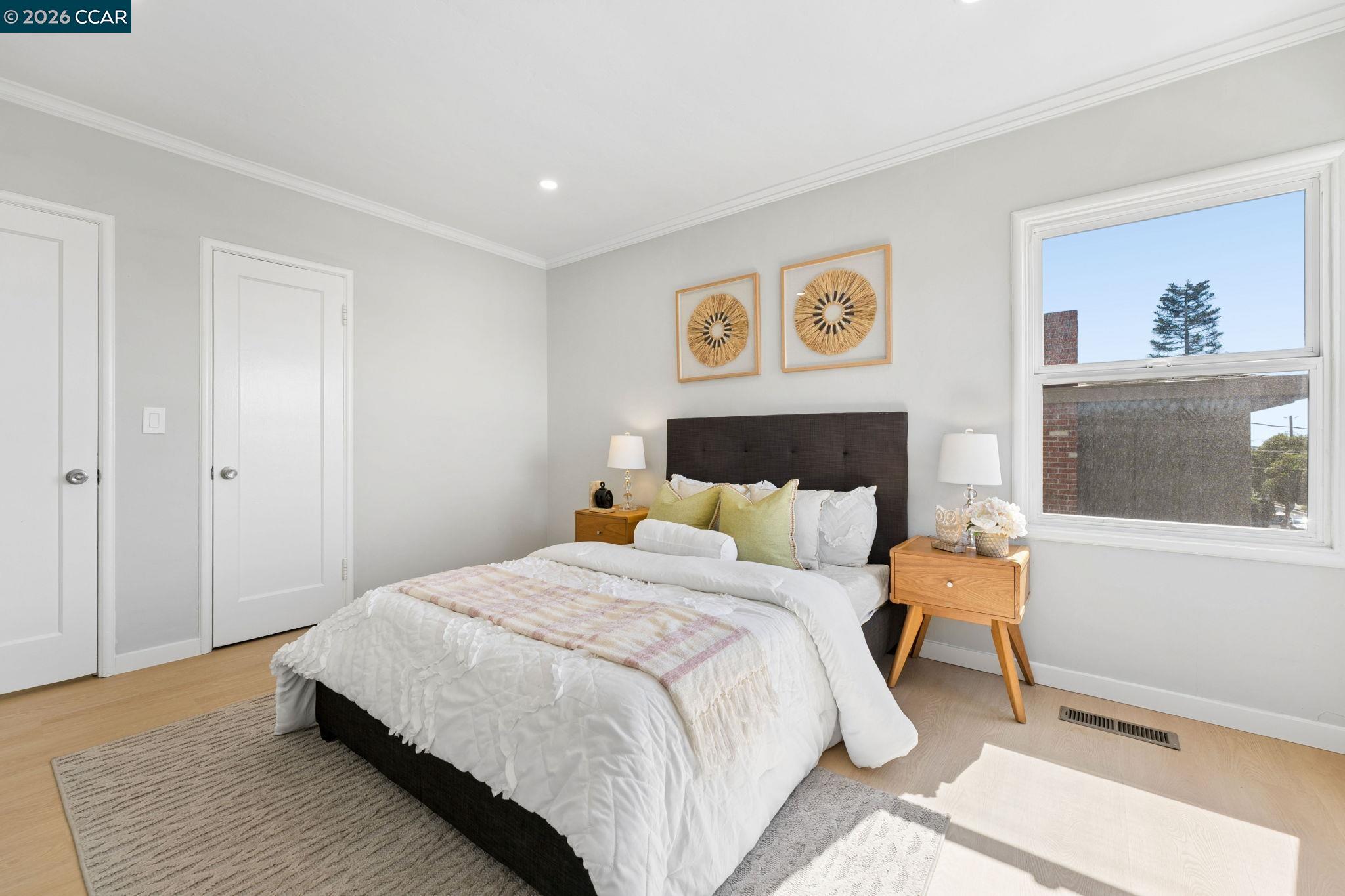 Staged bedroom with a dark upholstered bed, pale green pillows, wooden nightstands, and a large sunny window.