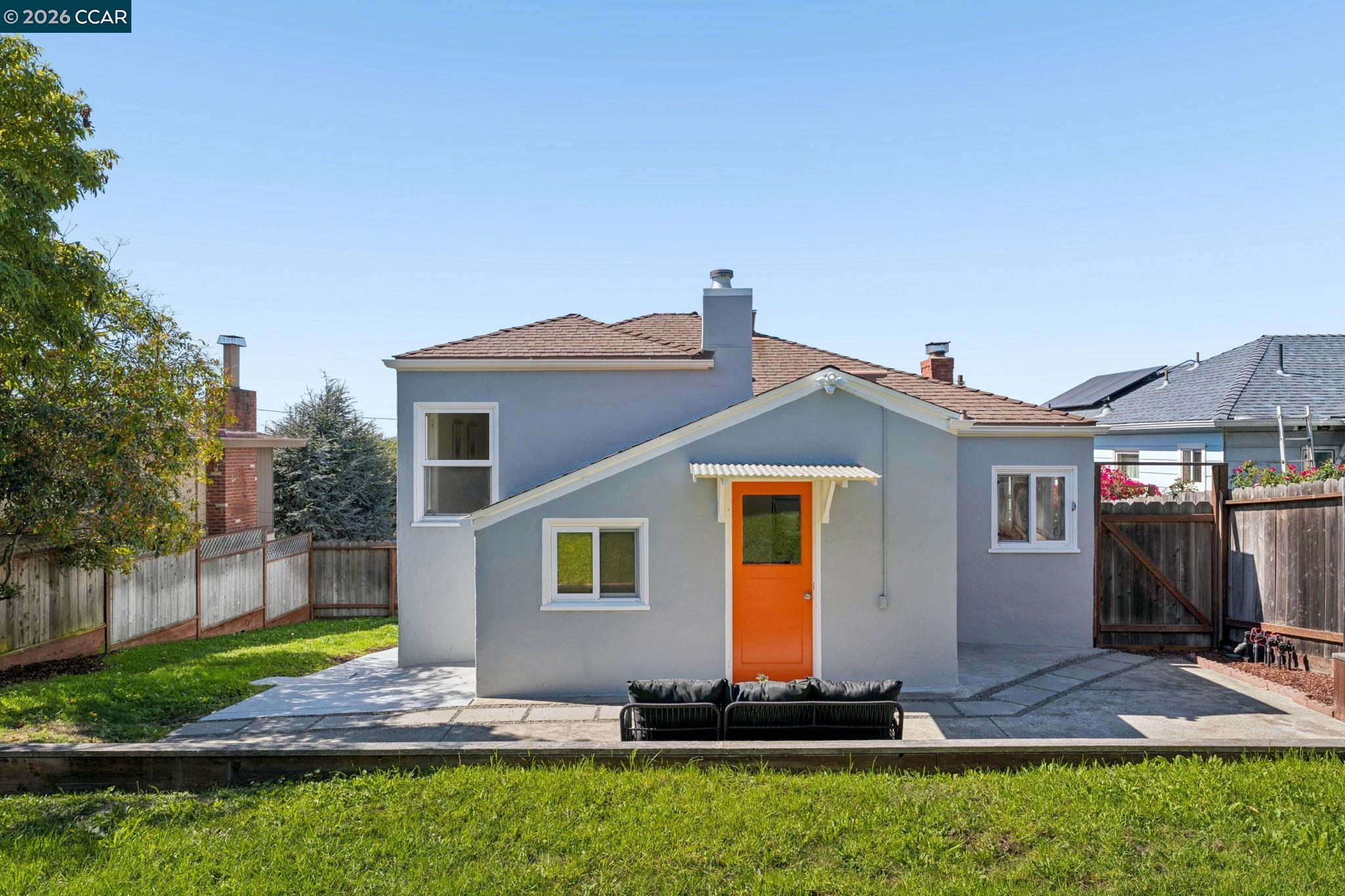 Gray house with a bright orange rear door, paved patio, outdoor sofa, and fenced lawn.