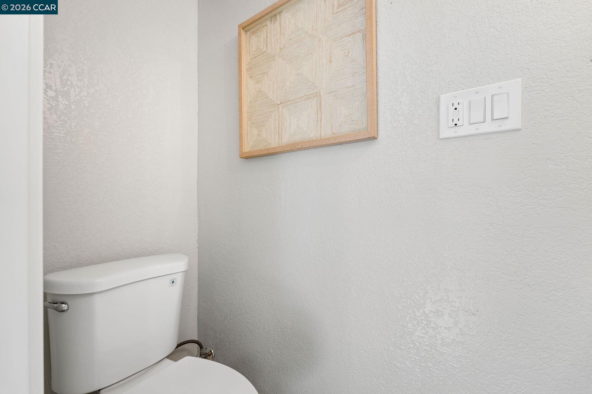 Bathroom toilet beneath framed geometric artwork, beside a wall plate with an outlet and switches.