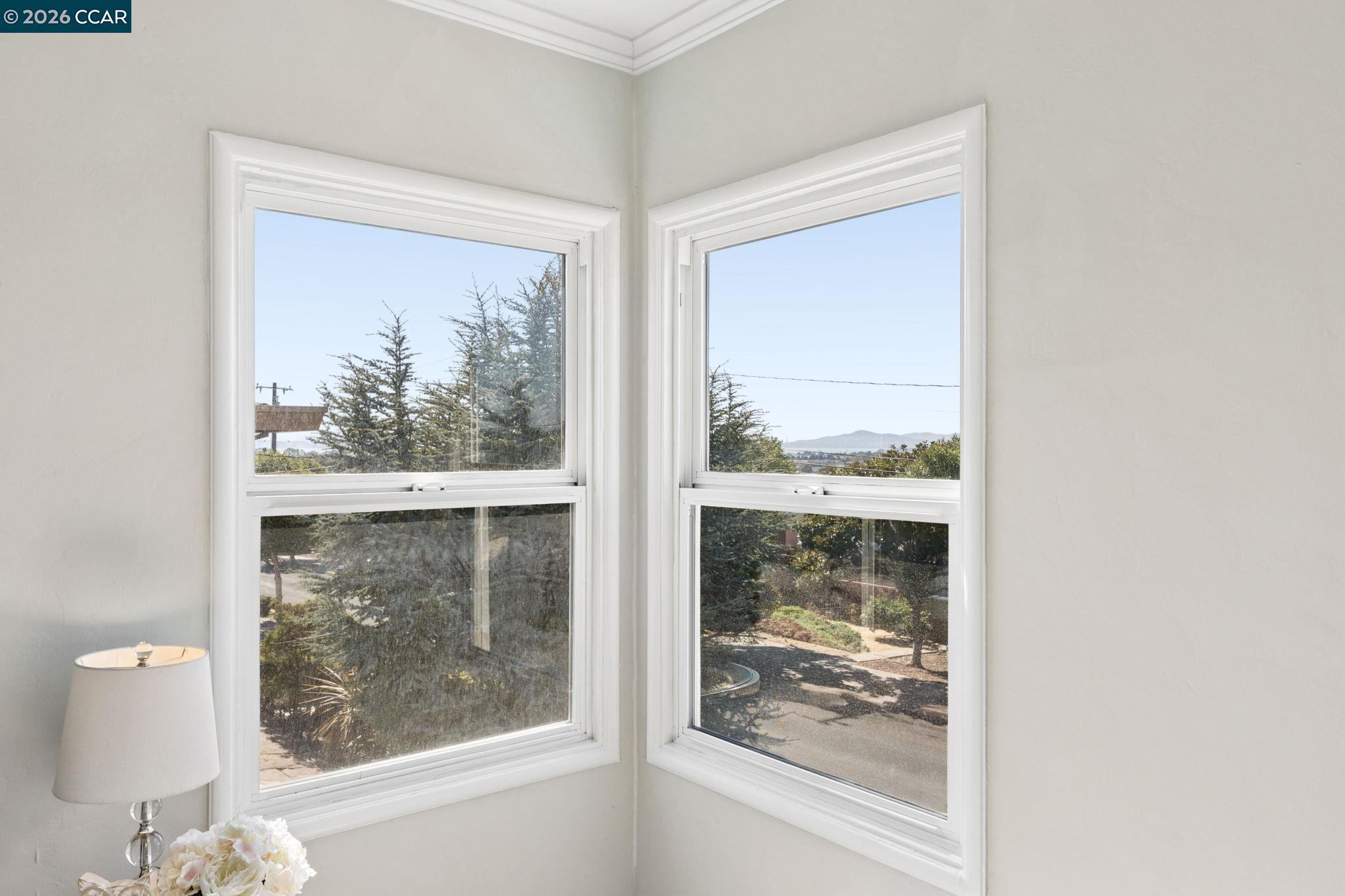 Bright room corner with two windows overlooking trees, a garden path, and distant hills.