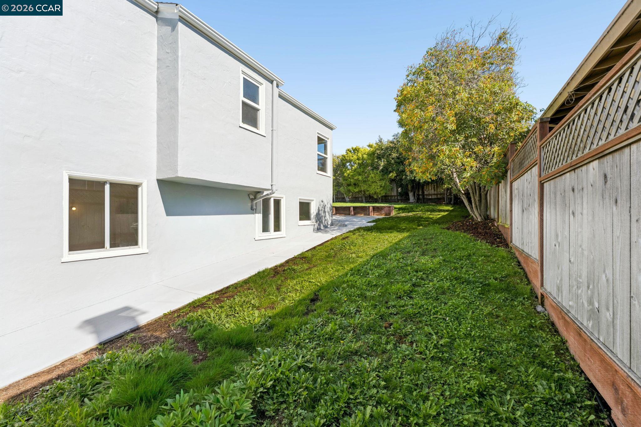 Side yard beside a white two-story house, with a grassy lawn, wooden fence, and paved patio.