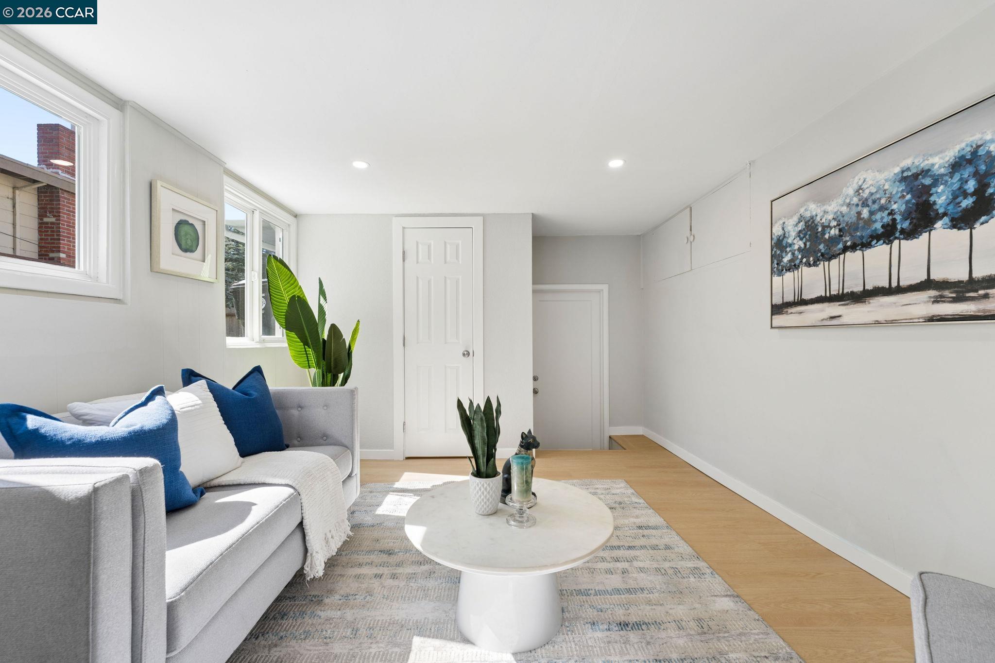 Bright living room with a gray sofa, blue cushions, round white coffee table, and large windows.