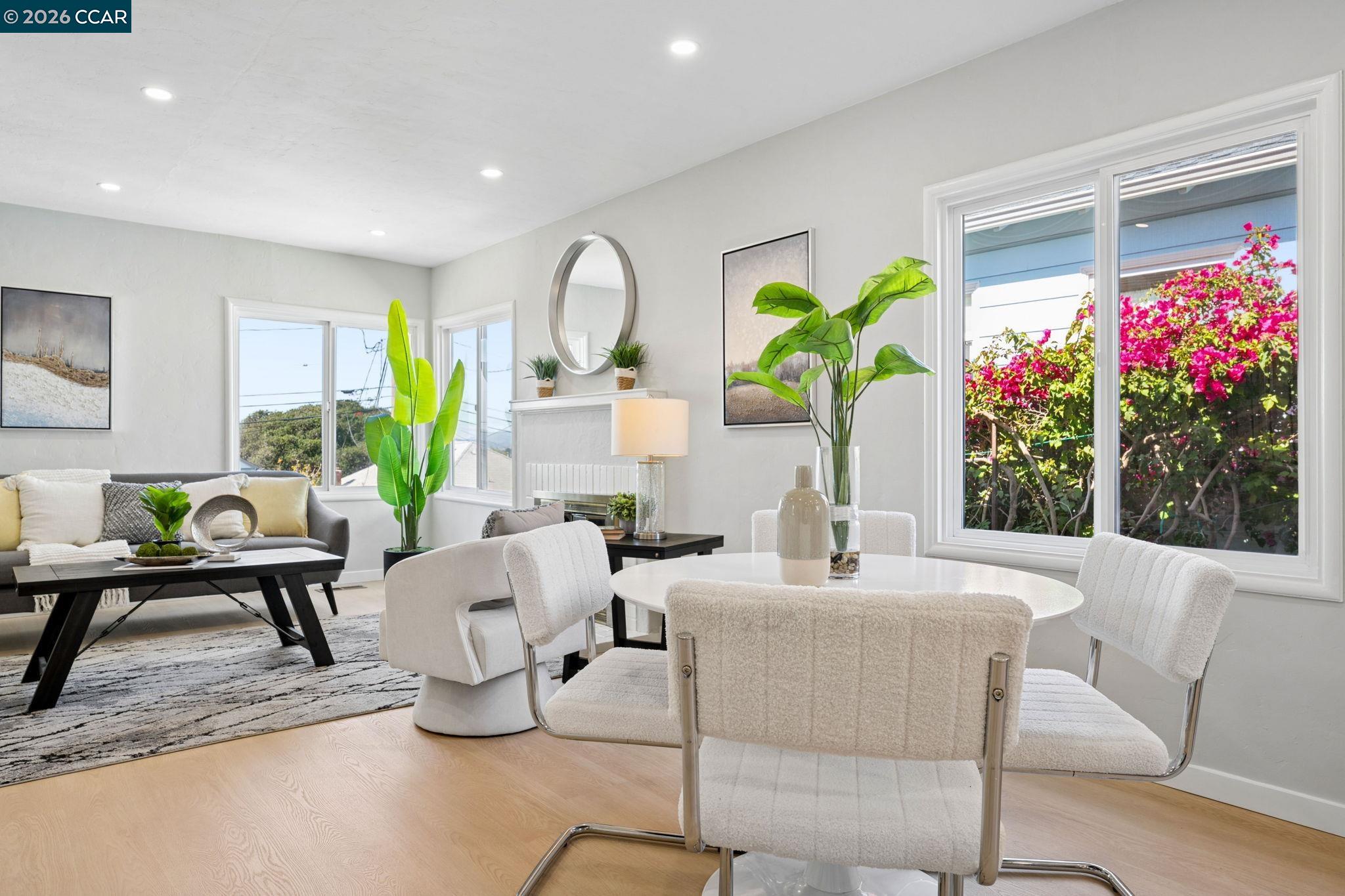Staged living and dining room with white furnishings and large windows overlooking pink flowers.