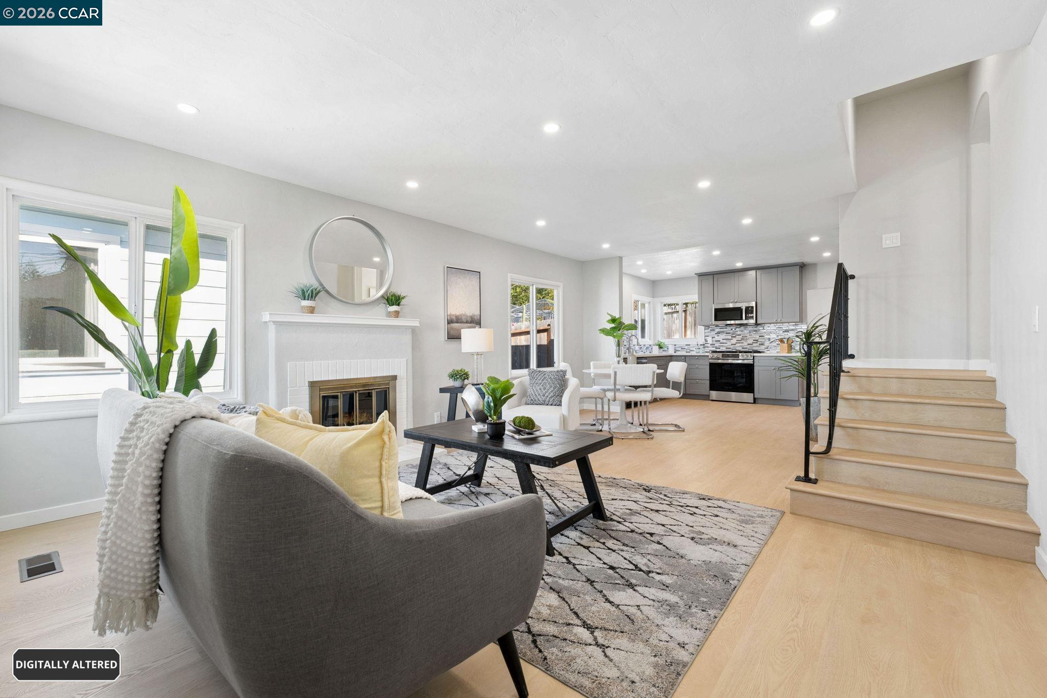 Staged open-plan living room and kitchen with a fireplace, gray sofa, gray cabinetry, and staircase.
