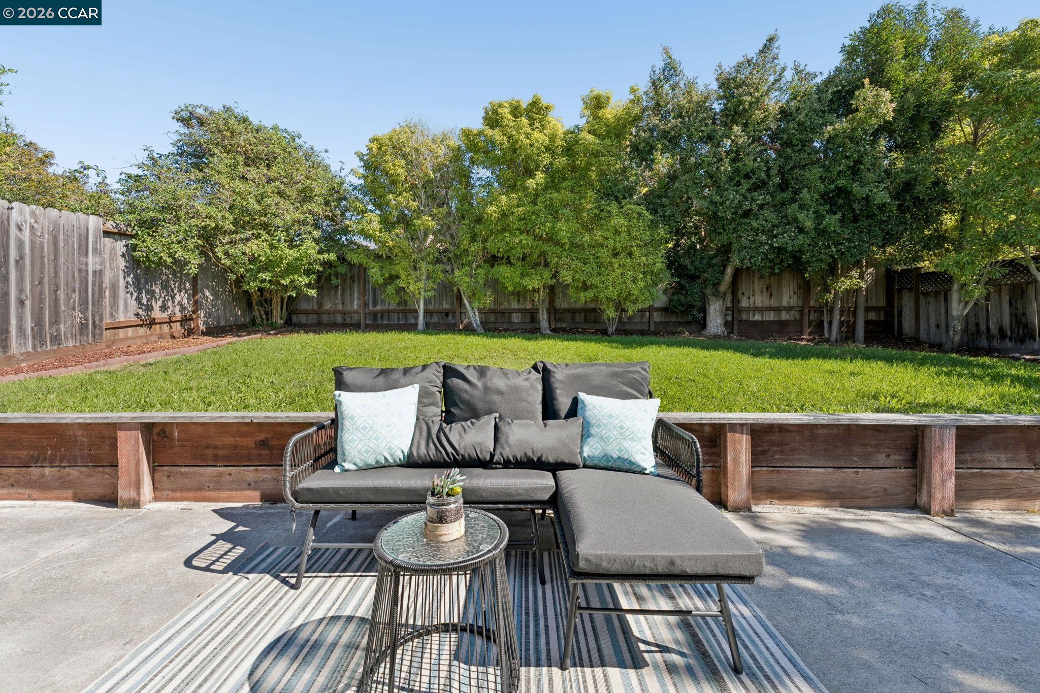 Backyard patio with gray chaise lounge seating beside a tree-lined lawn and wooden fence.