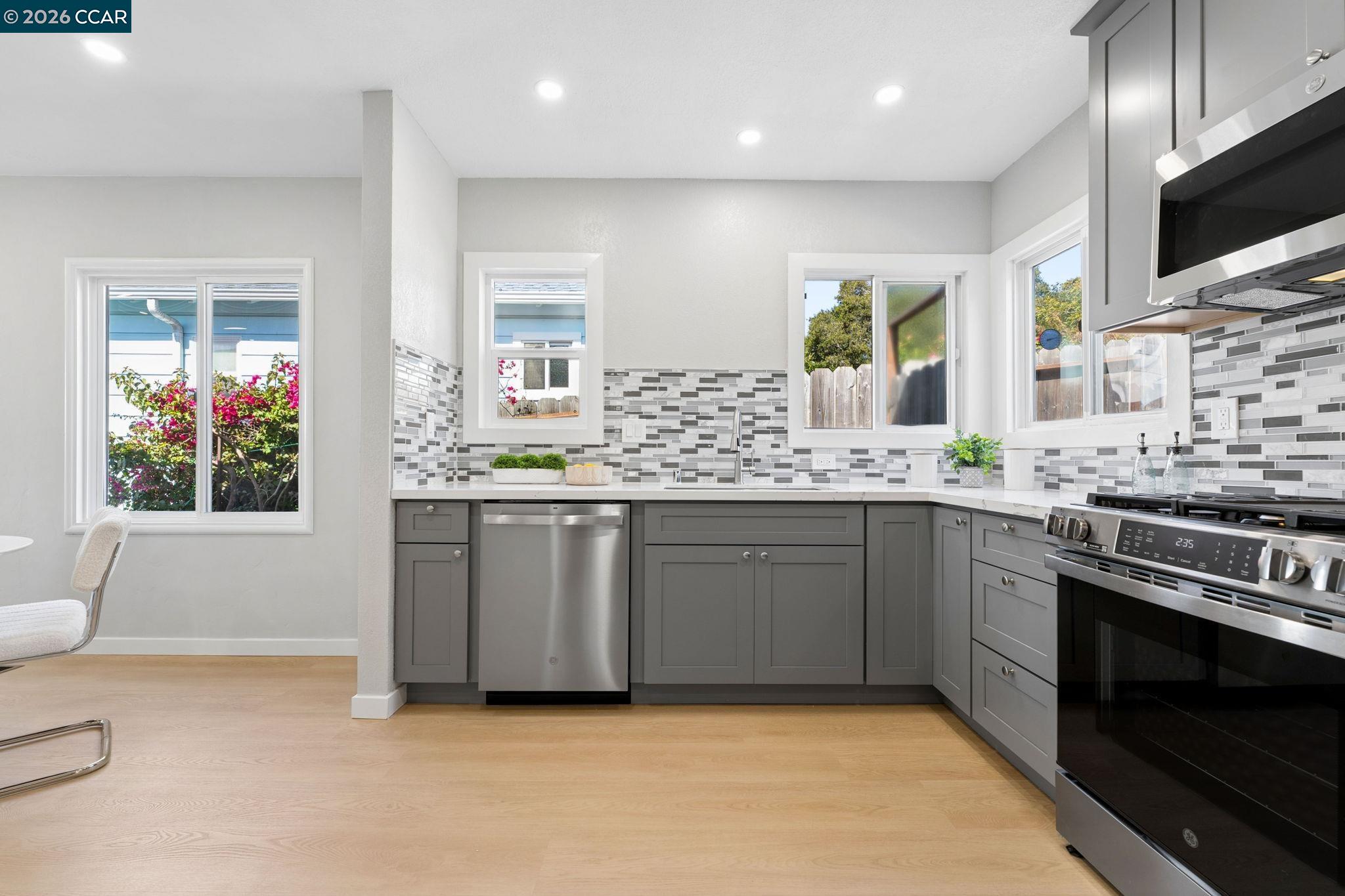 Bright kitchen with gray cabinets, stainless steel appliances, mosaic backsplash, and windows.